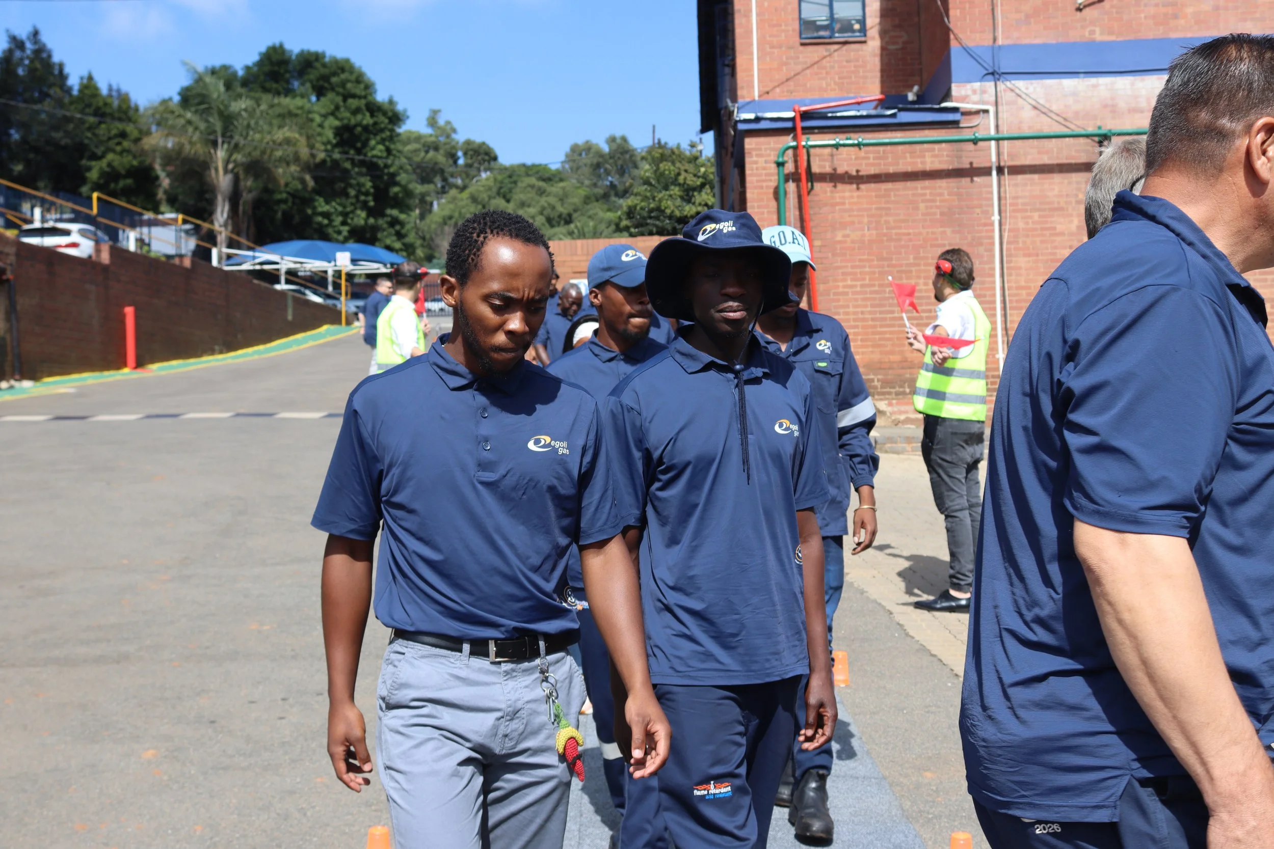 Group of people wearing blue uniforms, walking outdoors near a brick building, some with hats, on a sunny day.