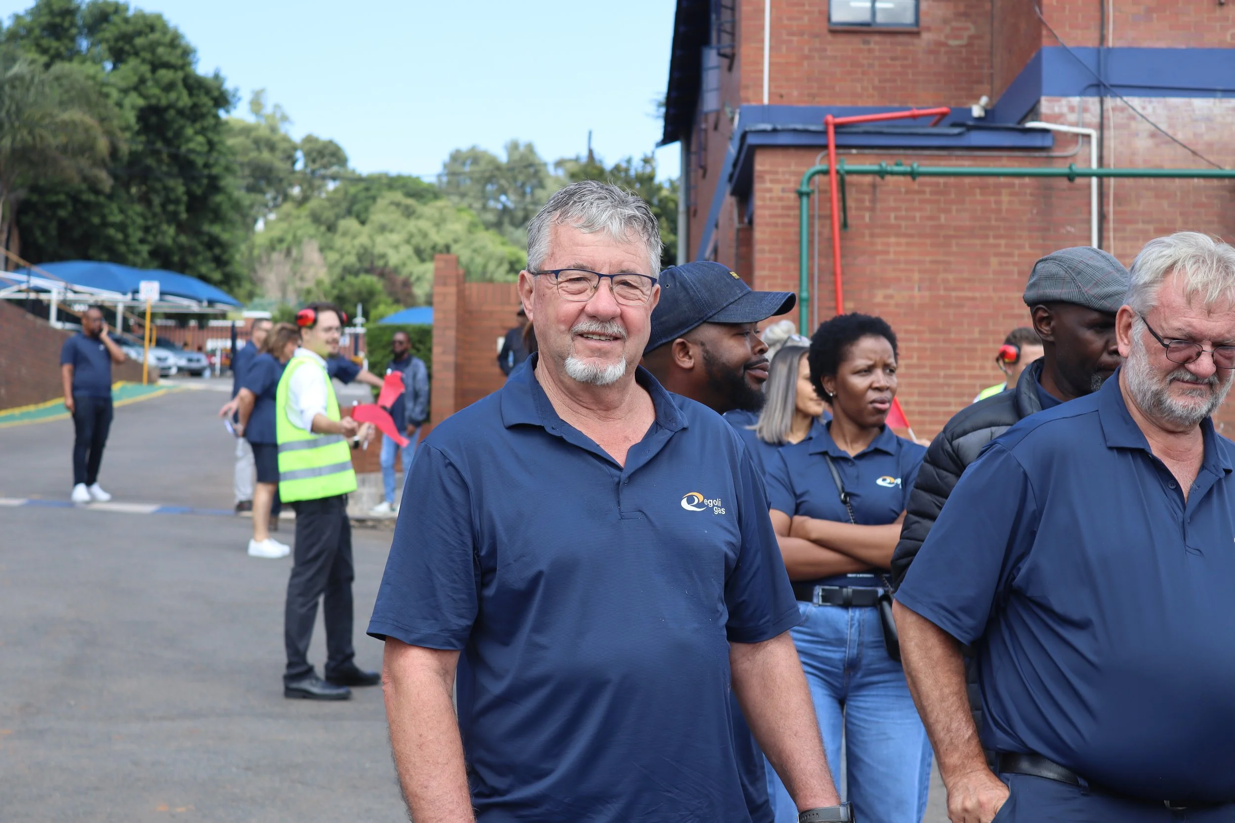Group of people, including a man with glasses and gray hair in the foreground, standing outside near a brick building and trees, wearing shirts with a logo, possibly at an outdoor event or gathering.