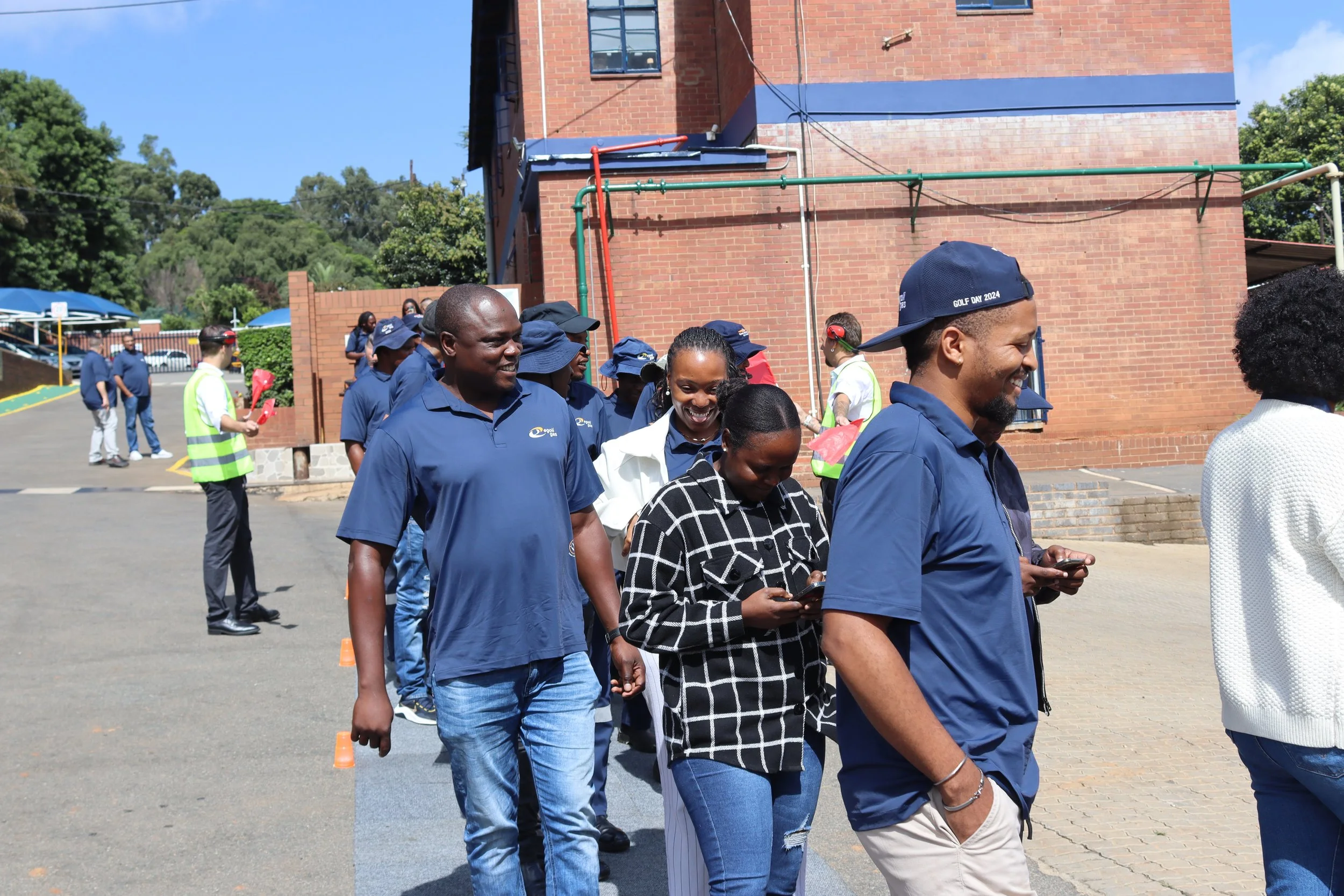 Group of diverse people standing in line outdoors on a sunny day, with a brick building and trees in the background, some wearing blue shirts and caps, some holding phones.