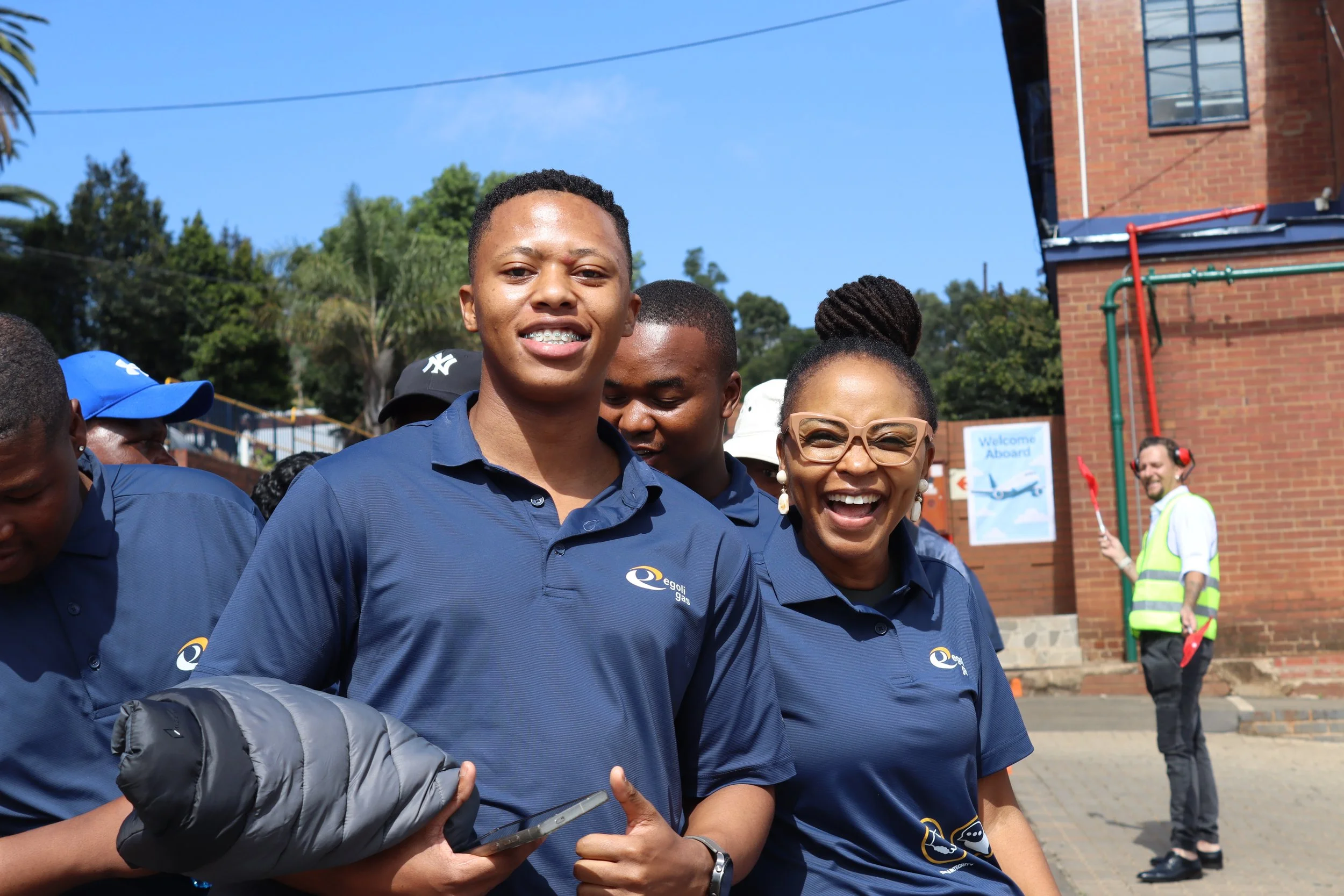 Group of smiling people wearing navy blue shirts at an outdoor event, with a brick building, trees, and a clear blue sky in the background.