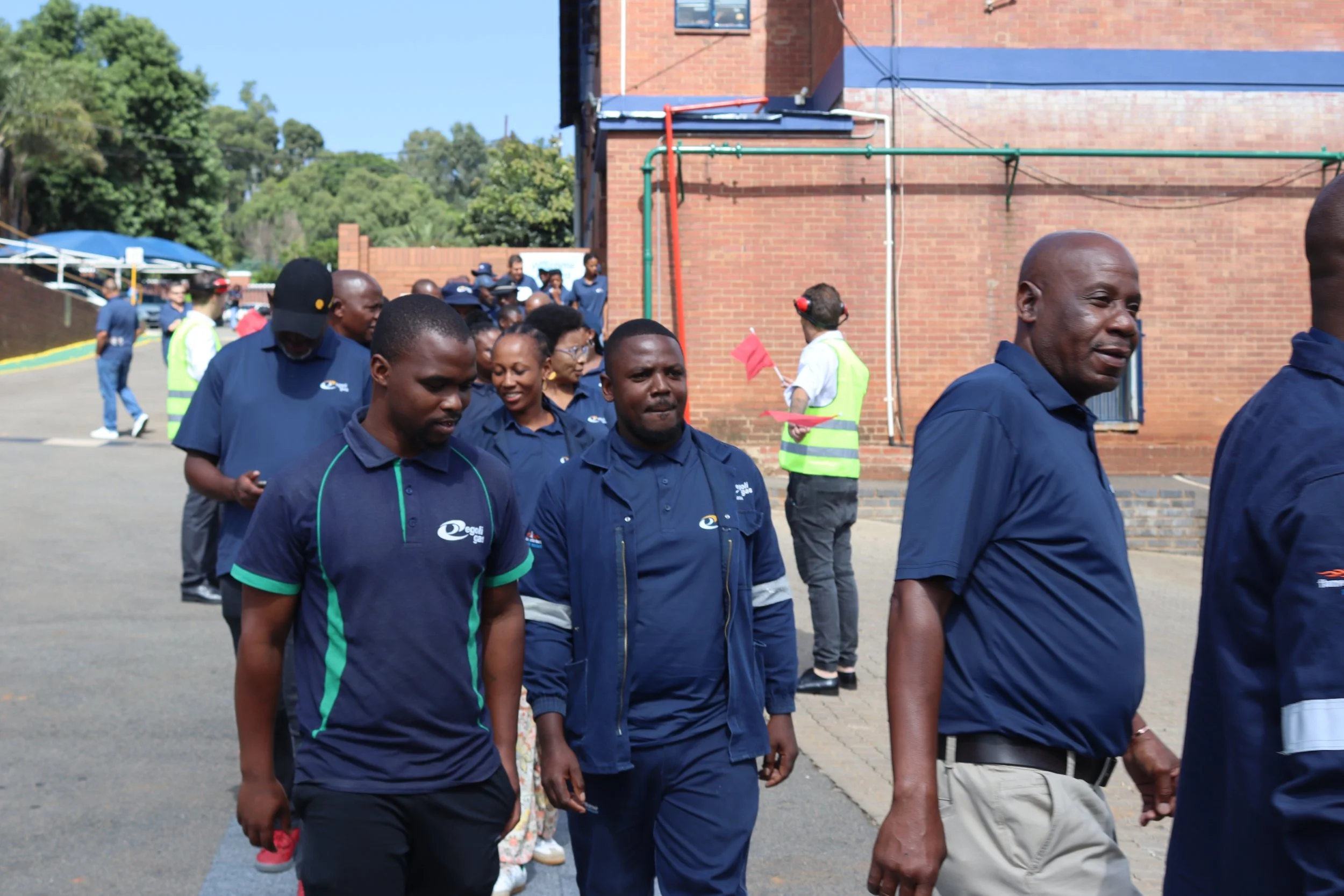 A group of people walking outdoors near a brick building with some greenery and trees in the background. The people are dressed in dark blue shirts, some with logos, and some are in casual attire. One person in a security vest is holding red flags.