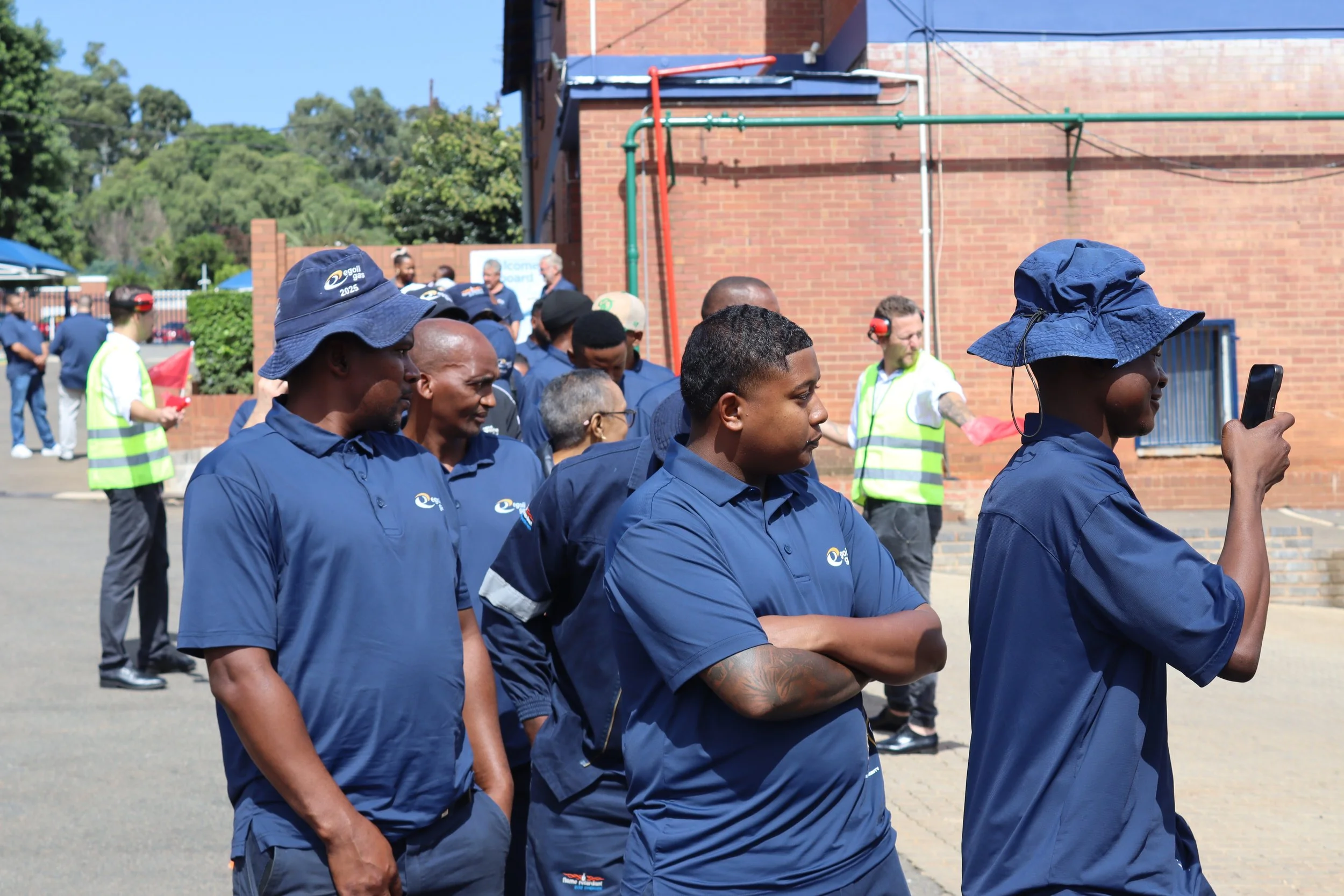 A group of people, mostly men and women, gathered outdoors near a brick building, some wearing blue uniforms with a logo, and a few wearing safety vests, during sunny weather.