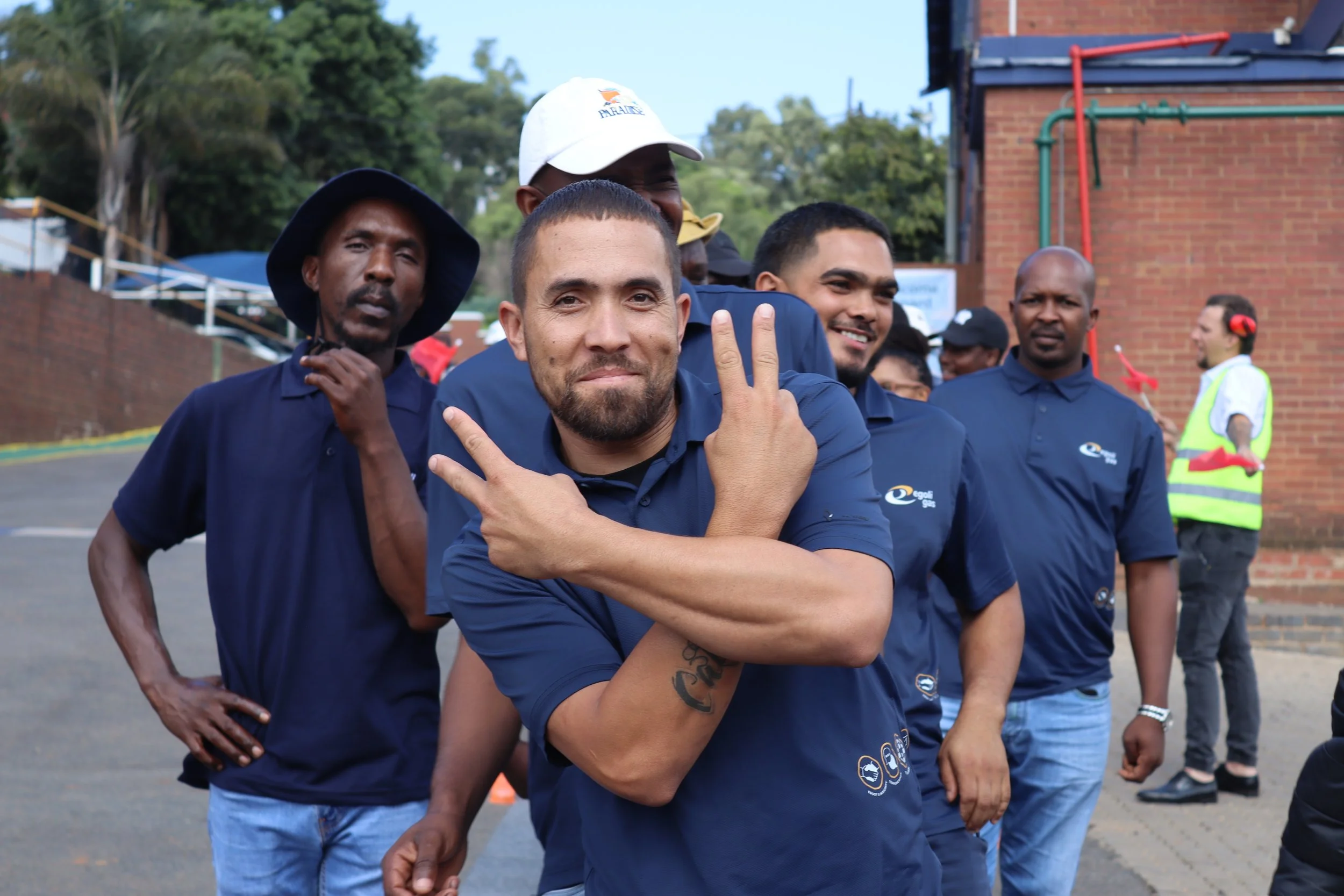 Group of men standing outdoors, with one in the foreground making a peace sign with his fingers, smiling. Others are standing behind him, some with serious expressions, with trees and a brick building in the background.