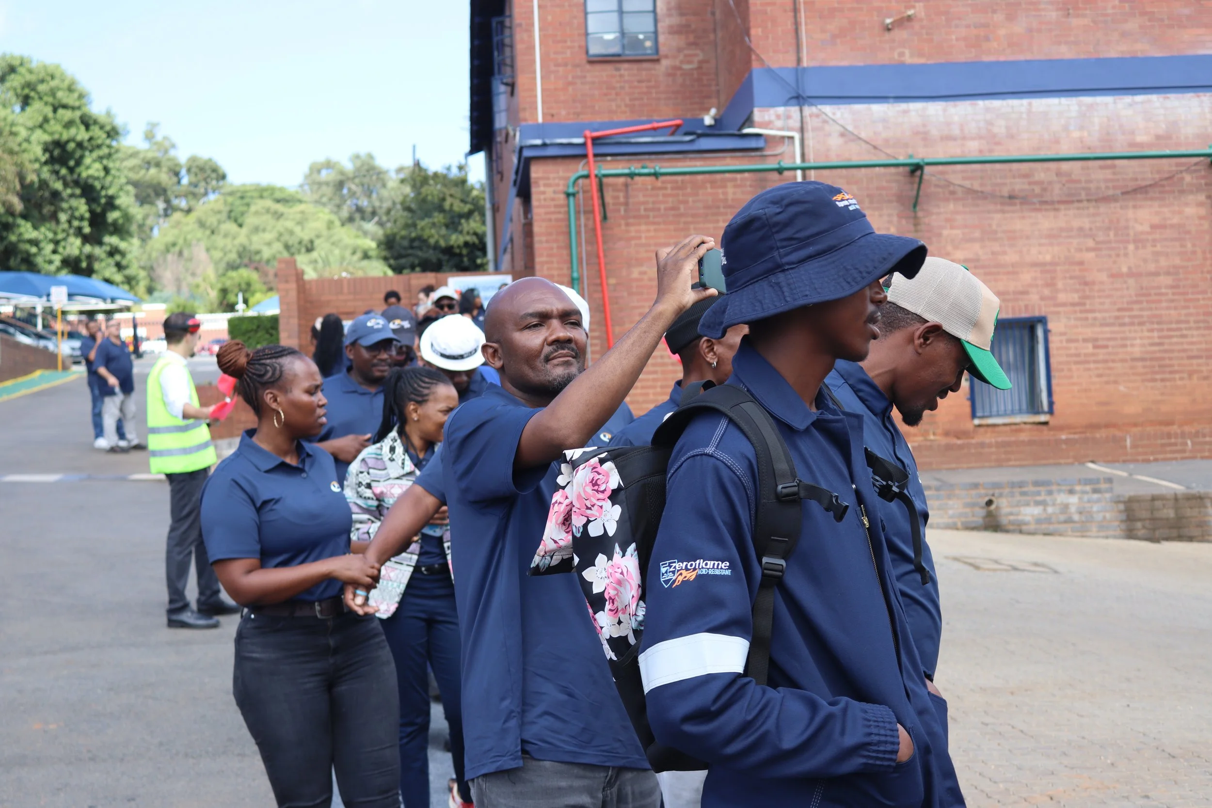 A group of diverse people standing in a line outdoors near a brick building. Some are looking down, while one man in front is taking a photo or video with a smartphone. Everyone appears to be engaged in a communal activity or event on a sunny day.
