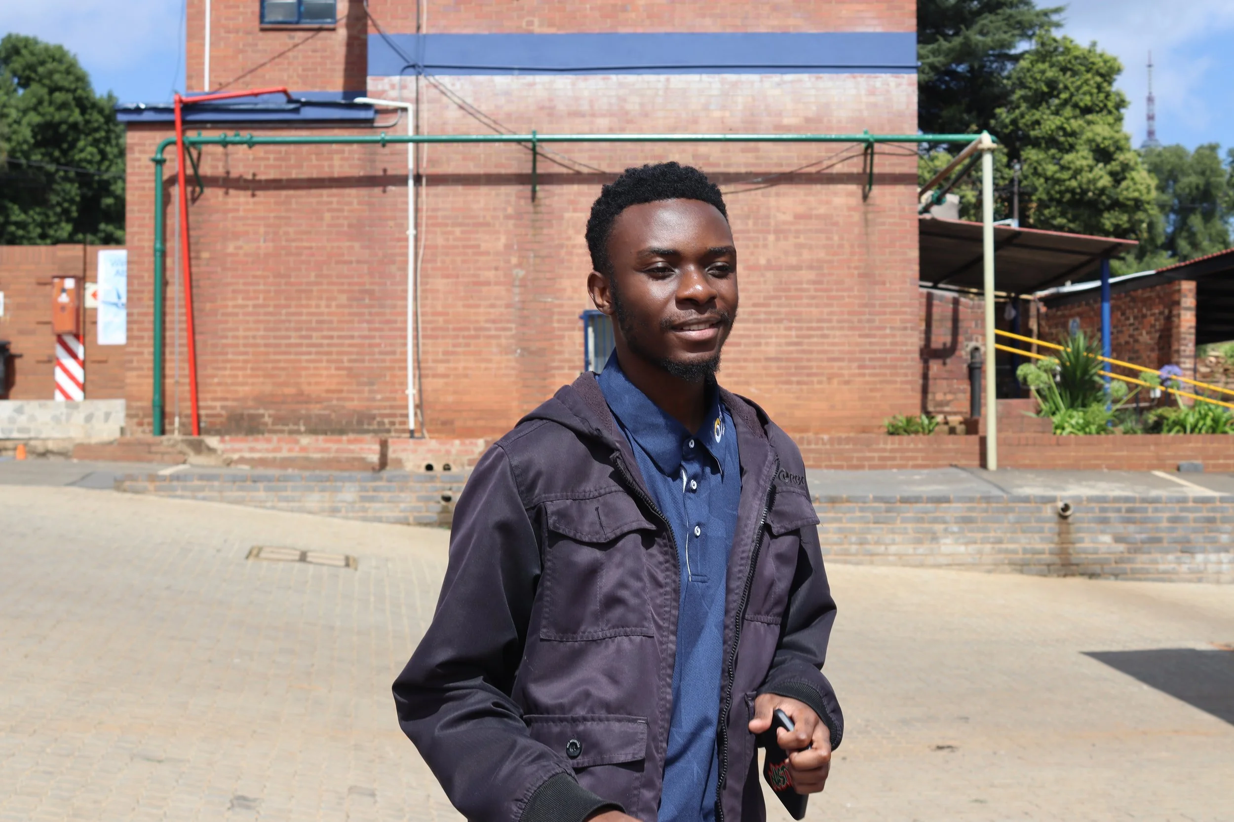 A young man with short curly black hair, a beard, wearing a dark jacket and a blue shirt, standing outside in front of a brick building with some greenery and a large antenna in the background.