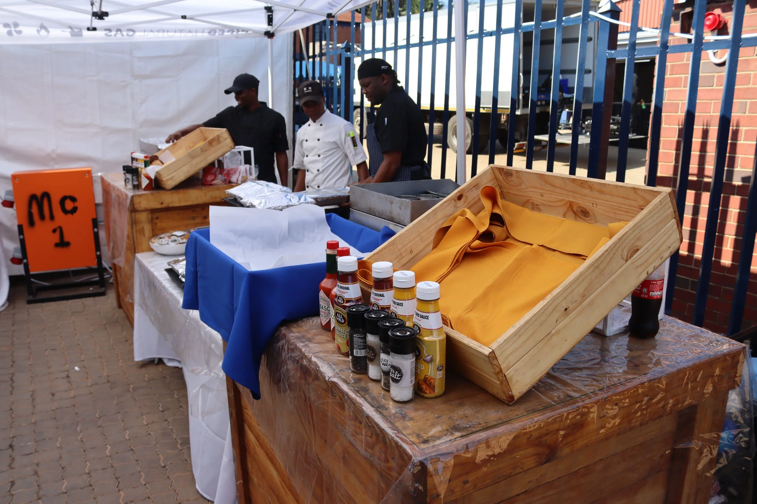 Food stall at outdoor event with three people preparing food, bottles of condiments on the table, and signs displaying prices.
