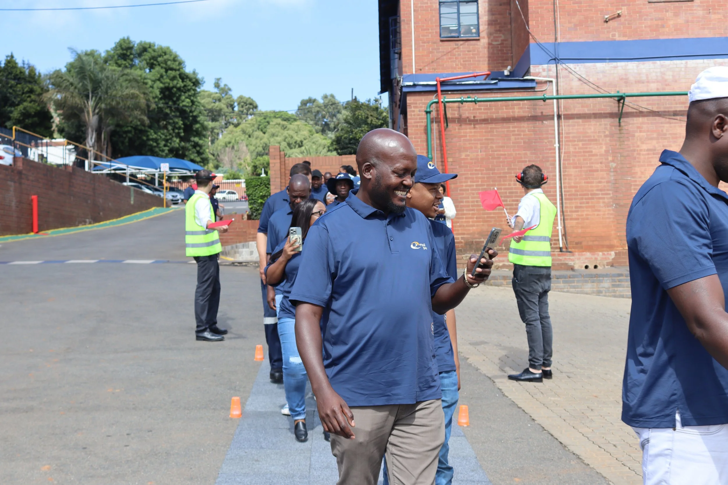 A group of people, dressed in blue shirts, walking and smiling outdoors near a brick building and trees, with some holding phones and others with safety vests and flags.