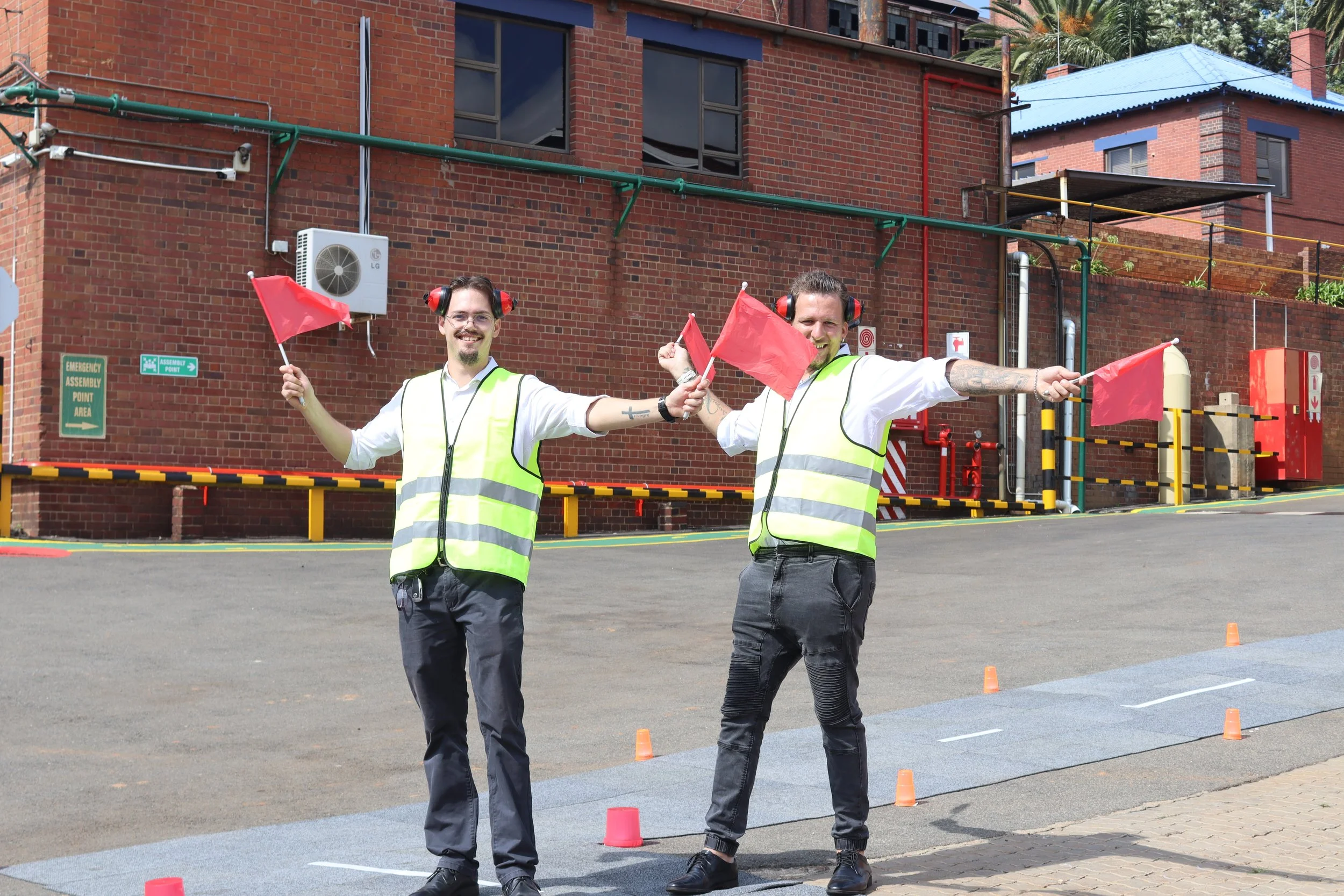 Two men wearing safety vests and earmuffs holding red flags, standing in front of a brick building with safety equipment and orange cones on the ground.