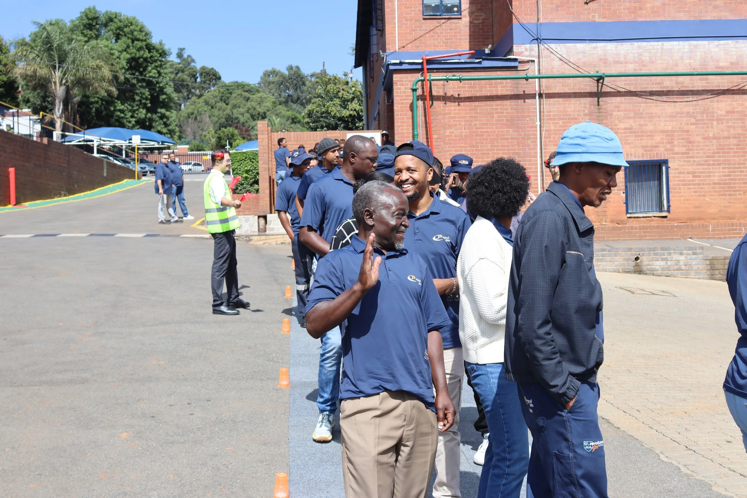 A group of diverse people standing in a line outdoors on a sunny day, some wearing blue shirts and hats, participating in an organized event near a brick building with trees in the background.