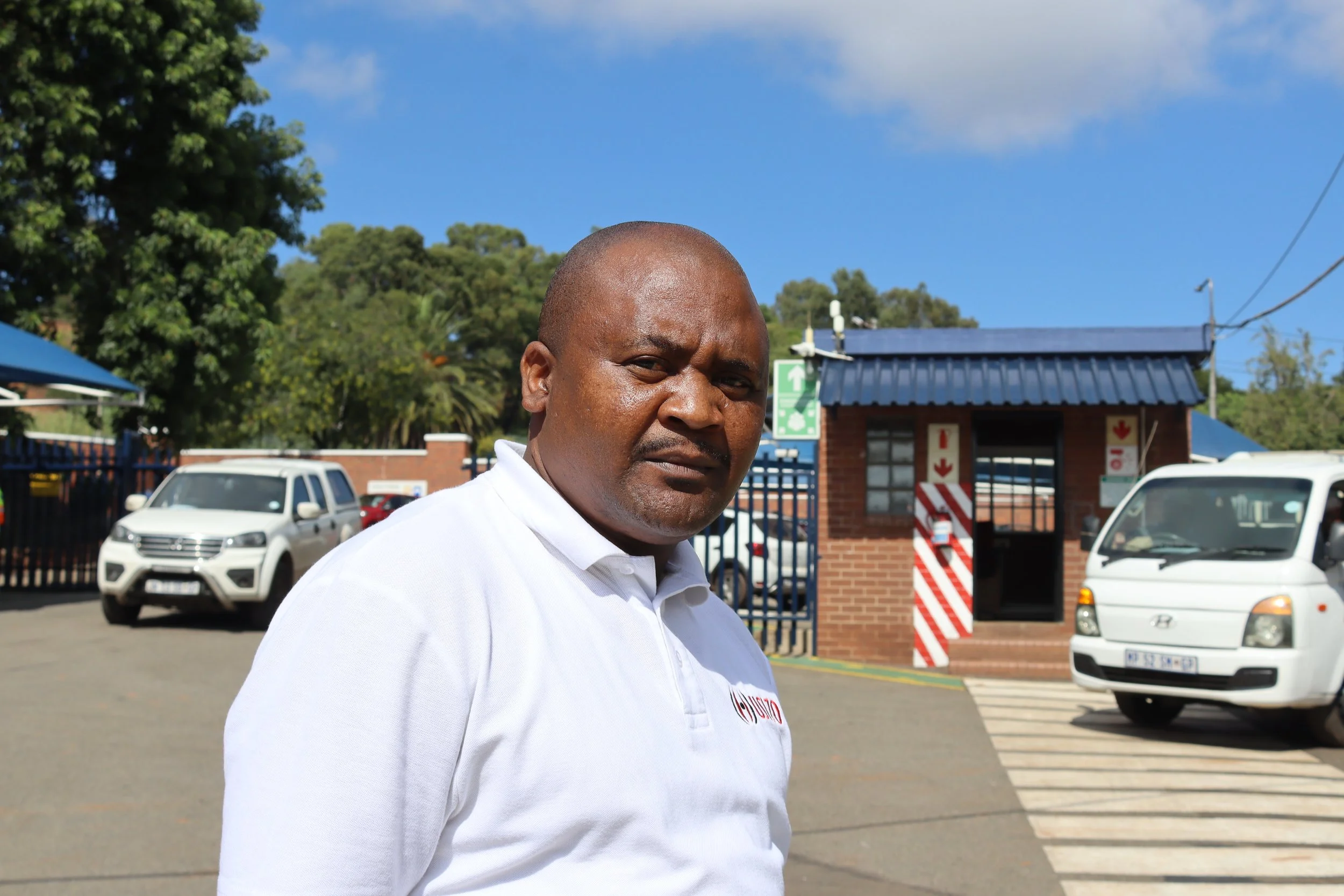 A middle-aged man with dark skin, a shaved head, and wearing a white shirt is standing outdoors near a parking lot with several vehicles. There are trees, a small building with a blue roof, and a blue sky in the background.