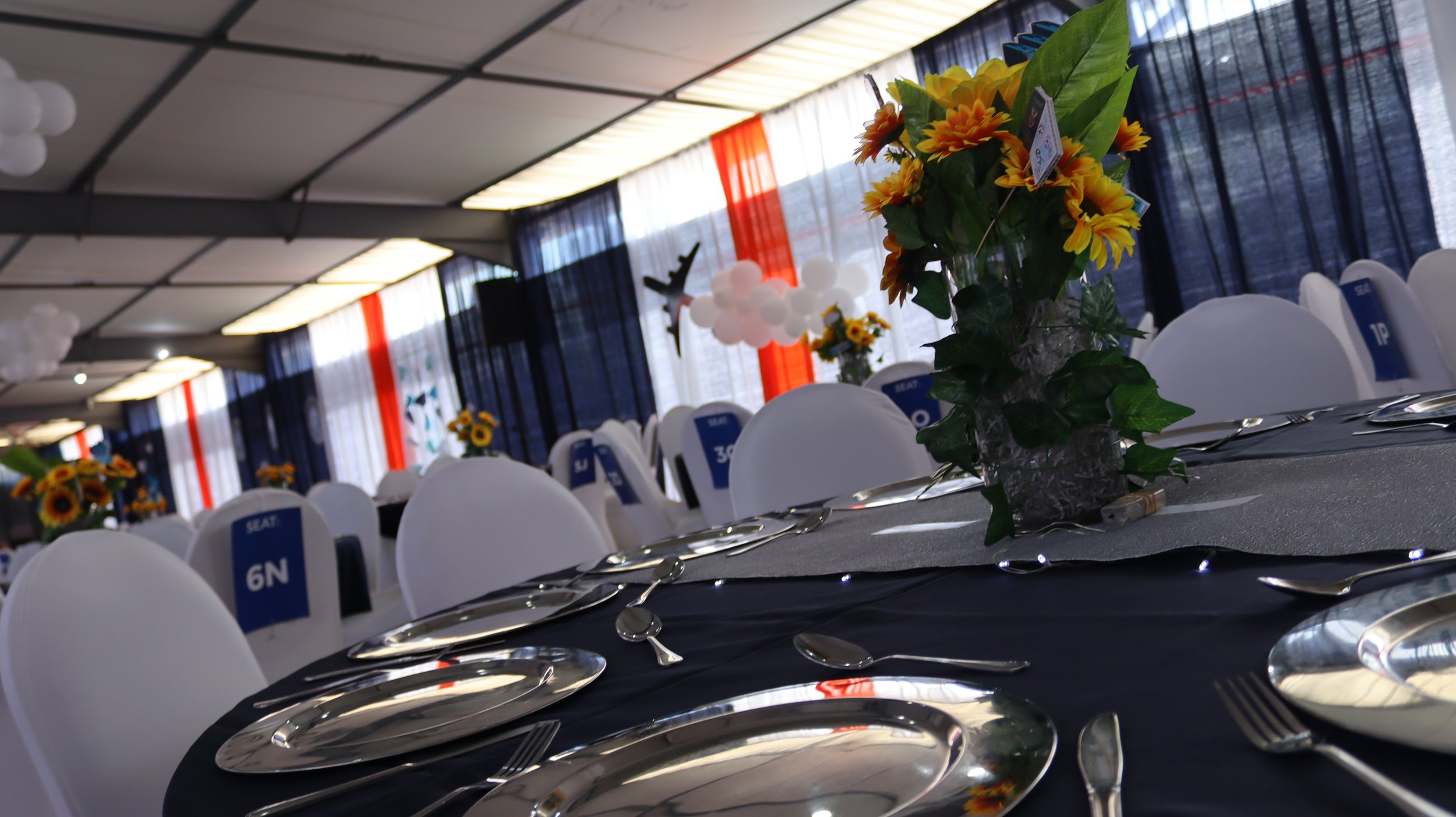 Event hall decorated with floral centerpieces featuring sunflowers, with white chairs and tables set for a formal dining arrangement, black tablecloths, and blue, white, and orange fabric draped along the windowed ceiling.