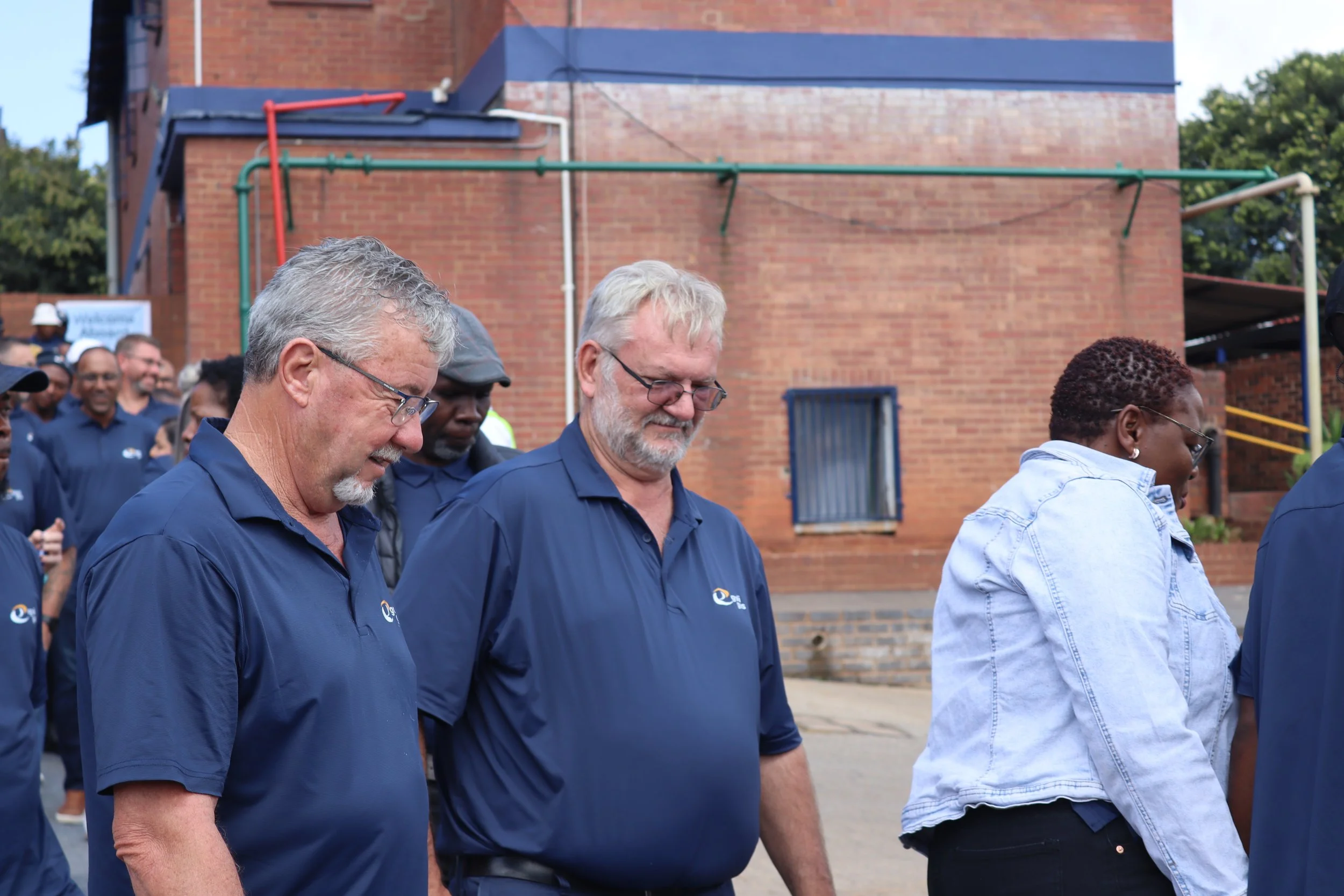 A group of people walking outdoors, dressed in blue shirts, with some looking downward and others looking ahead, near a brick building with pipes and trees in the background.