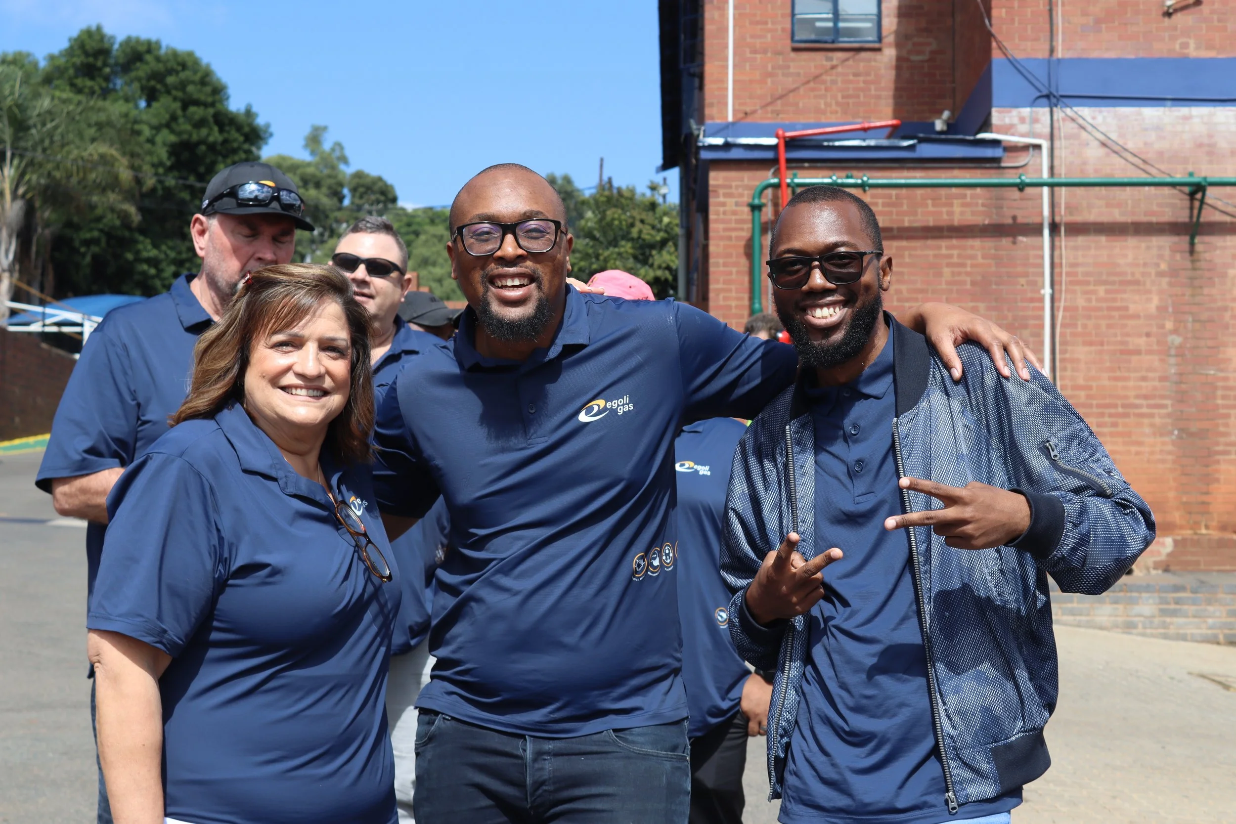 Group of smiling people wearing matching blue shirts with a logo, standing outdoors in front of a brick building and green trees.