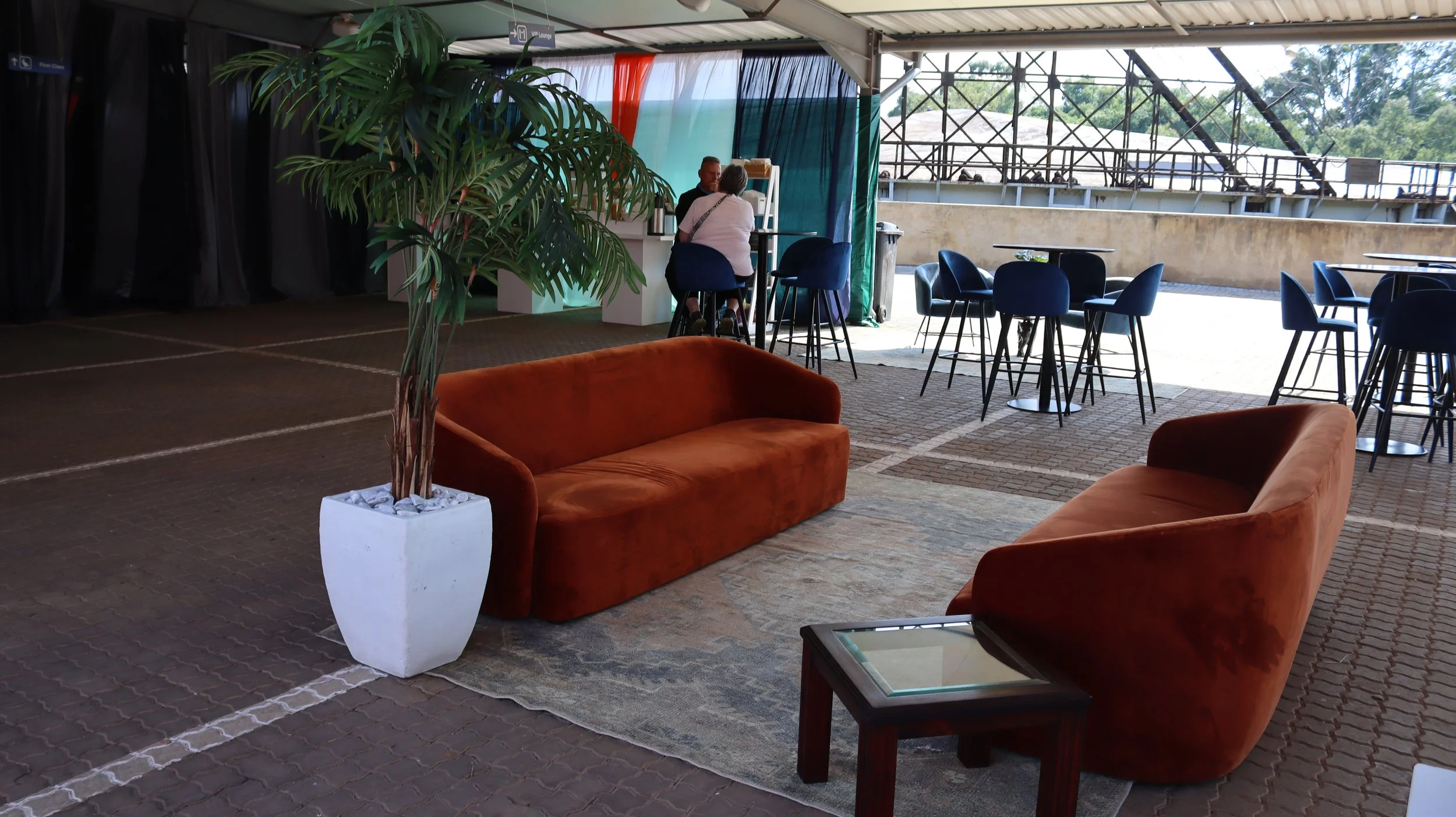 Indoor lounge area with orange velvet sofas, large potted plant, high tables with blue chairs, and a woman sitting with a man, with an outdoor view of a construction scaffold or framework in the background.