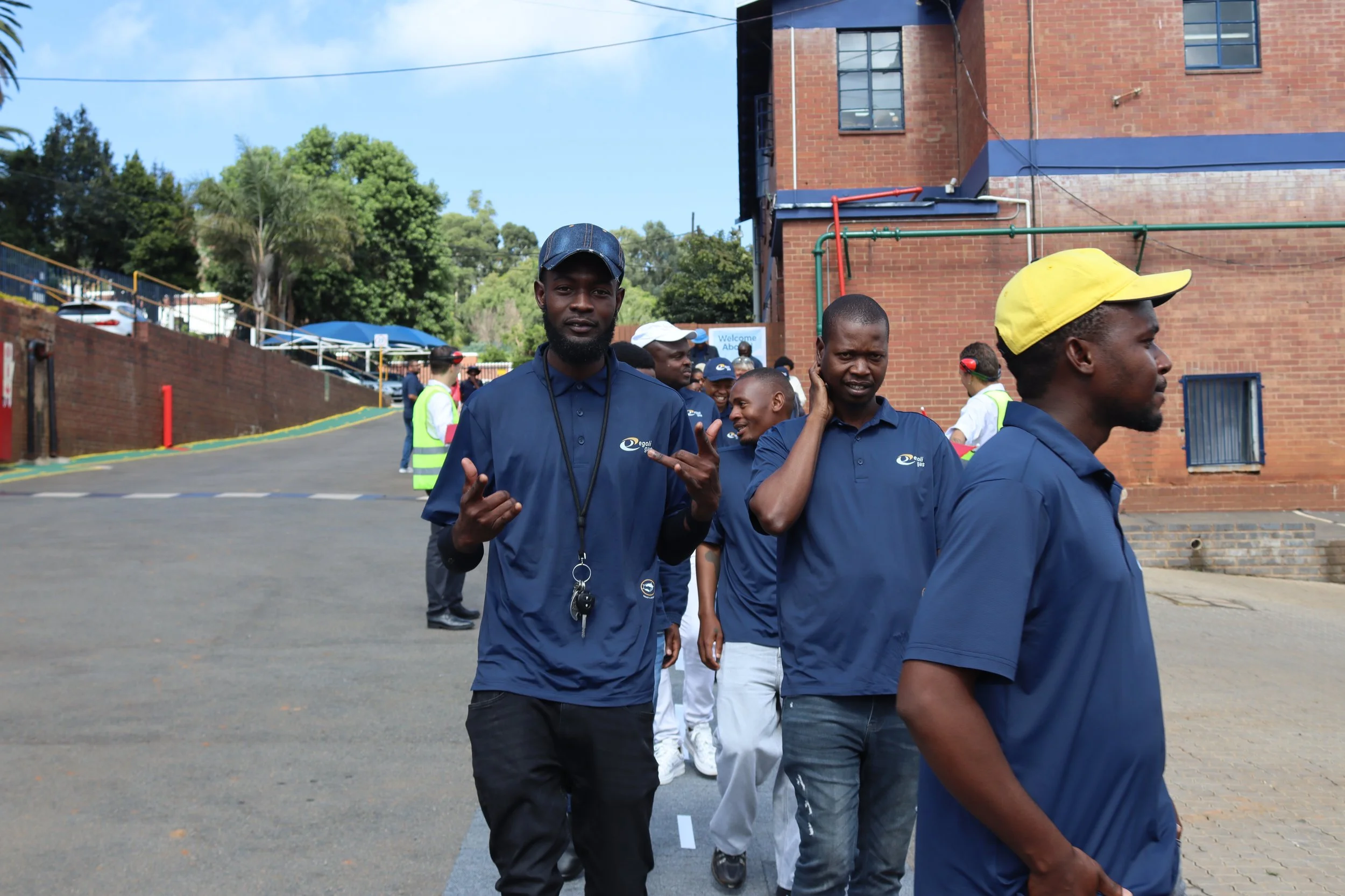 Group of young men walking outdoors near a red brick building, some talking and smiling, wearing dark blue shirts with a logo, with a background of trees and a blue sky.