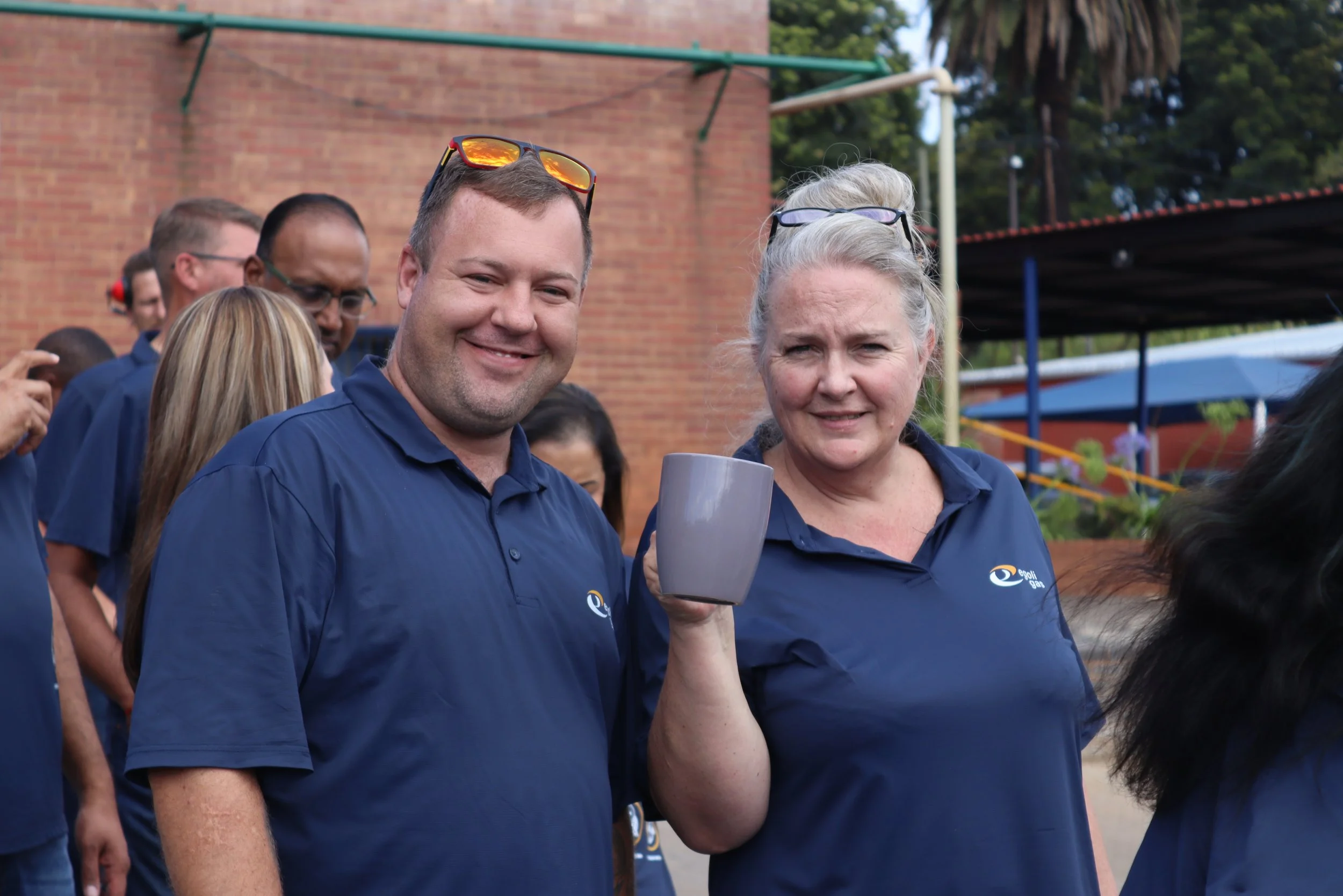 A group of people wearing matching dark blue shirts at an outdoor event, with two people in the foreground smiling at the camera. The man has sunglasses on his head, and the woman is holding a gray cup. In the background, there are additional people,