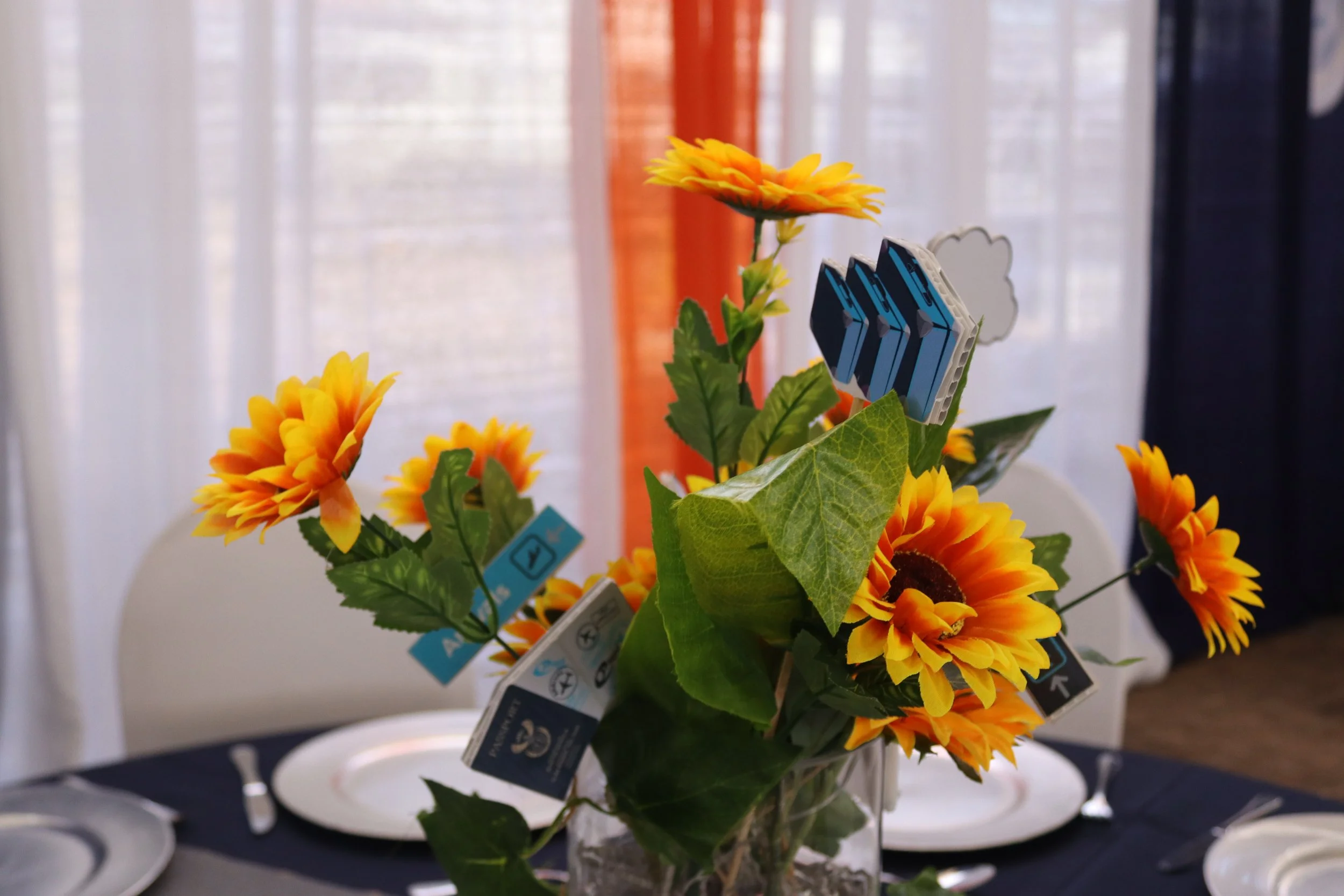 A small bouquet of artificial sunflowers with green leaves in a white vase, displayed on a table with white plates around it.