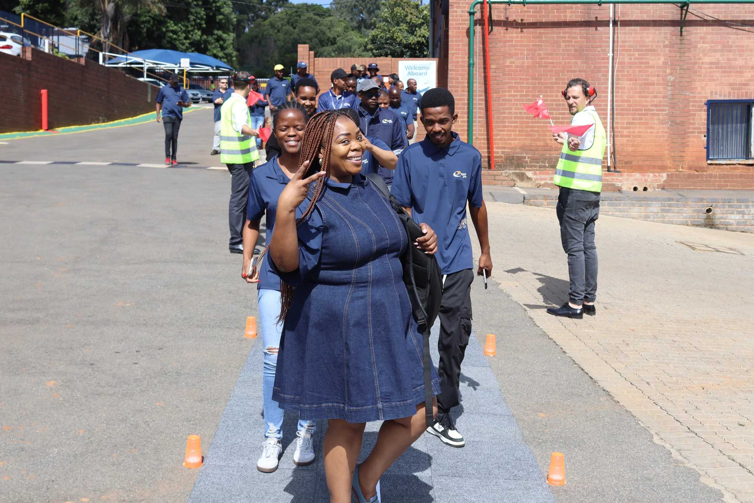 A group of people participating in an outdoor event, standing in line, with some holding red flags, near a brick building on a sunny day.