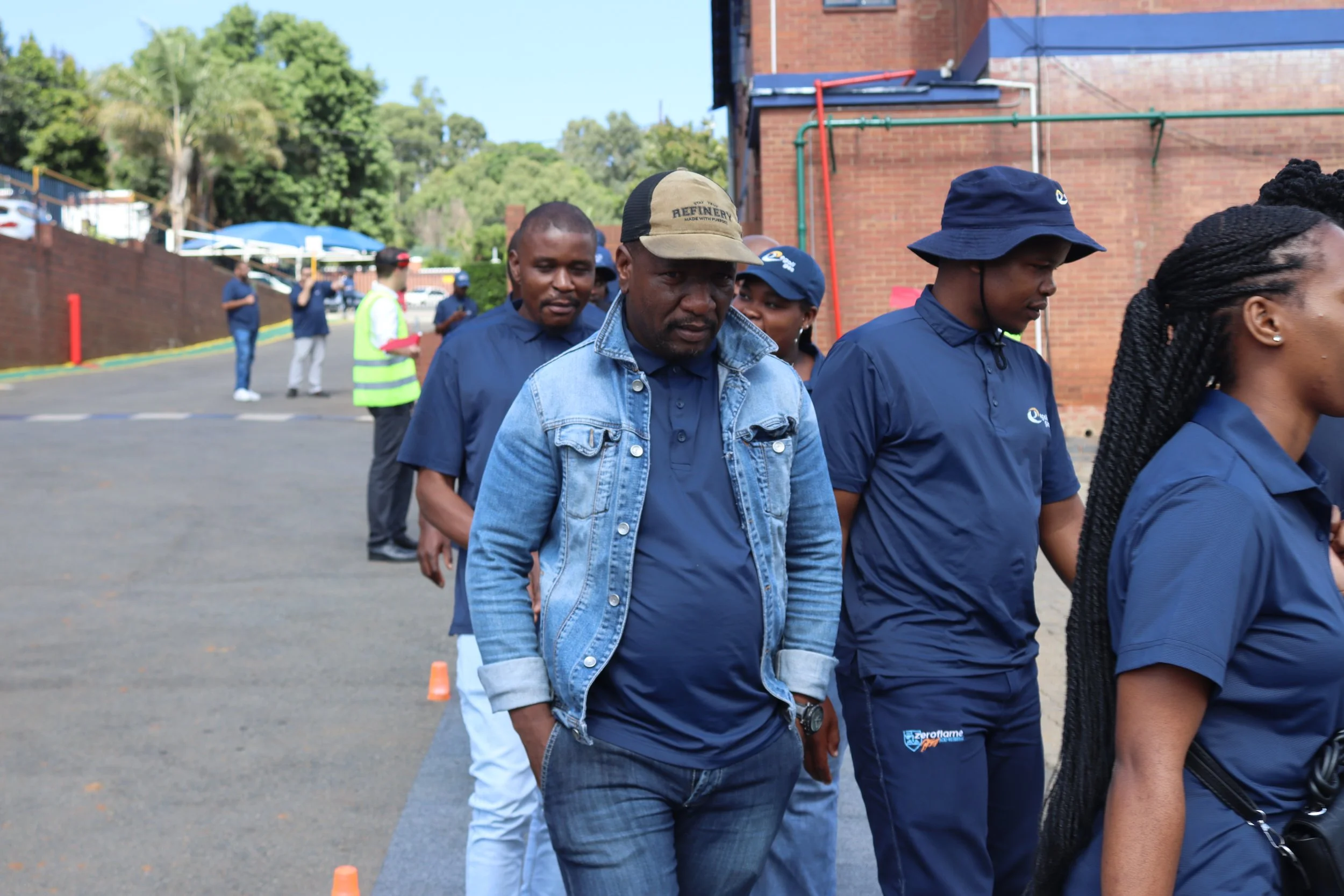 A group of people, including a man in a denim jacket and a woman with braided hair, walking outdoors near a brick building with trees in the background. There are other people standing behind them, some wearing high-visibility vests.