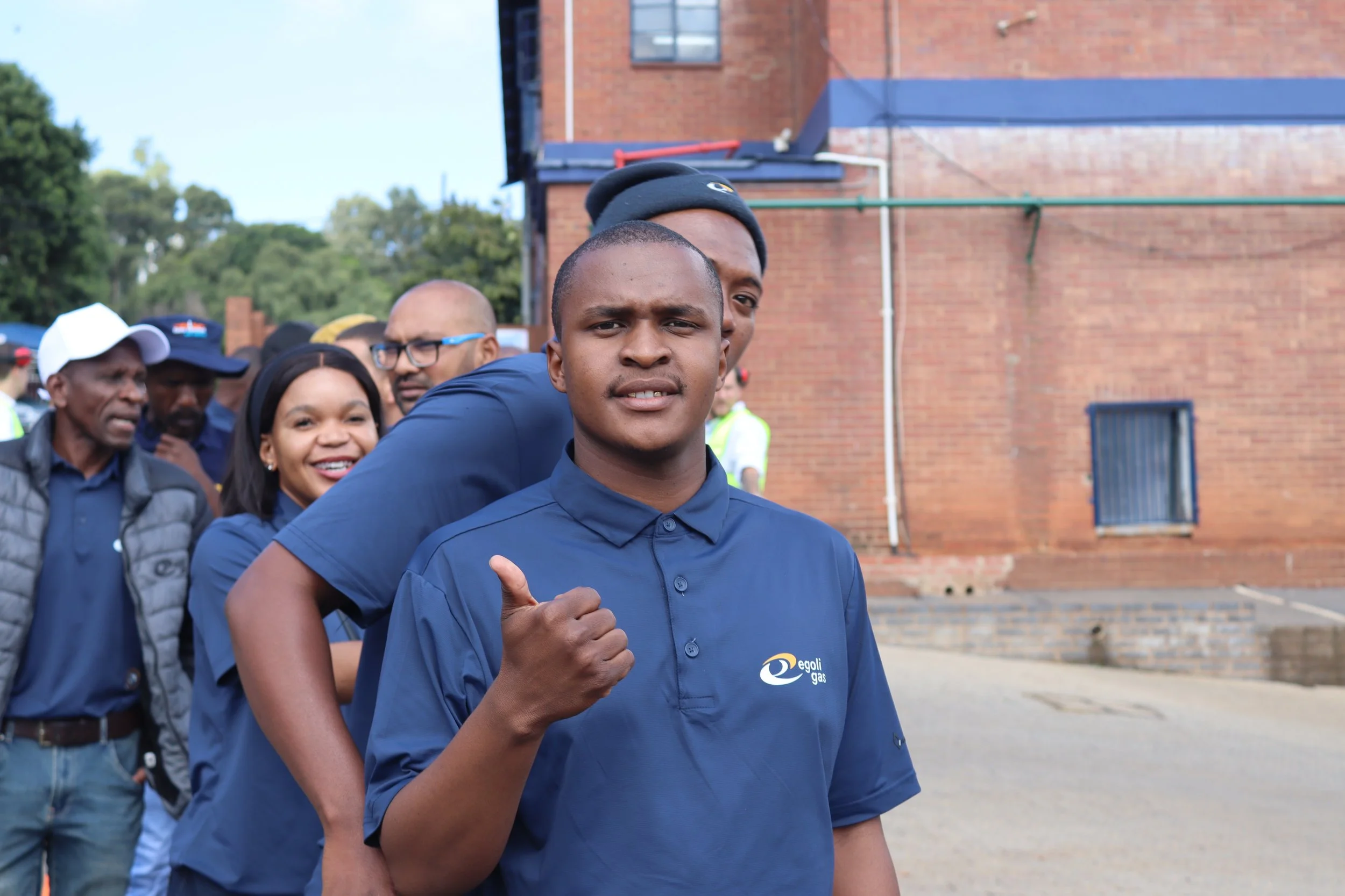 Young man in blue uniform with logo, giving a thumbs-up in a line of diverse people outdoors in front of a brick building and greenery, during daytime.