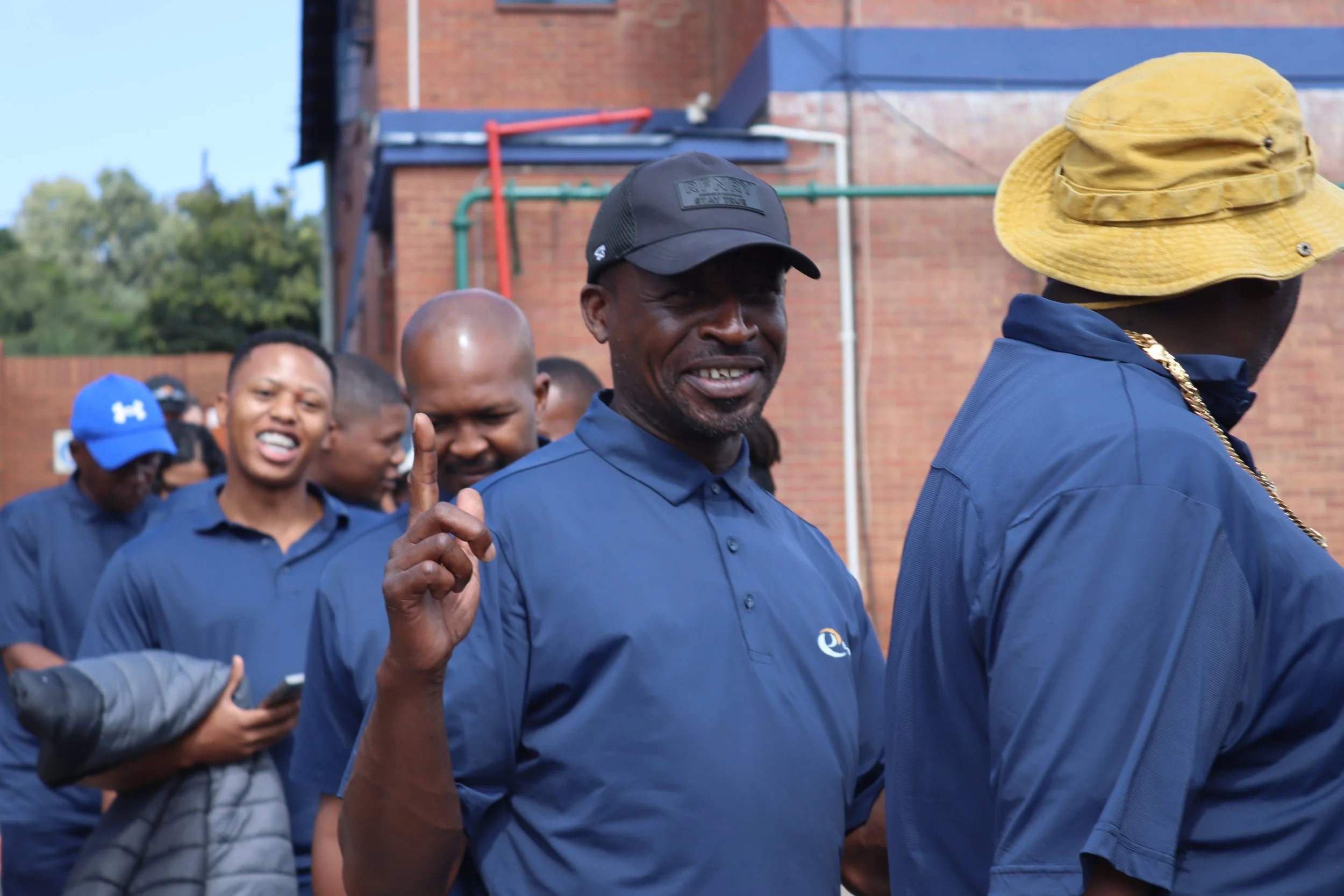 A group of people outdoors, some smiling and looking at their phones, wearing blue shirts, with a man in the foreground wearing a black cap and another wearing a yellow hat, in front of a brick building with colorful pipes.