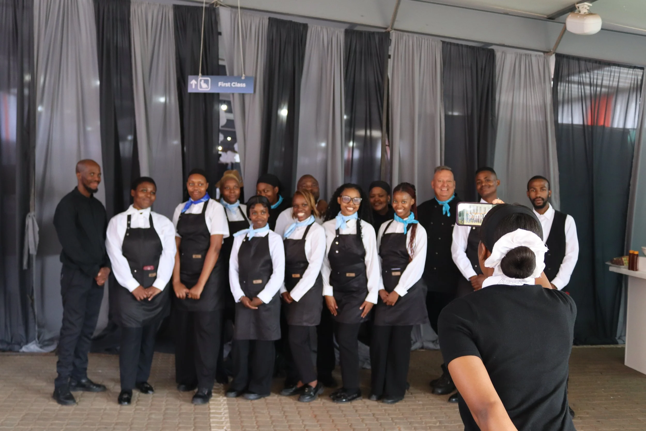 Group of people, including men, women, and children, dressed in black and white chef uniforms, standing together for a photo in front of curtains. Some are smiling, and a woman with glasses is in the center. A sign above indicates the area is for "Fi