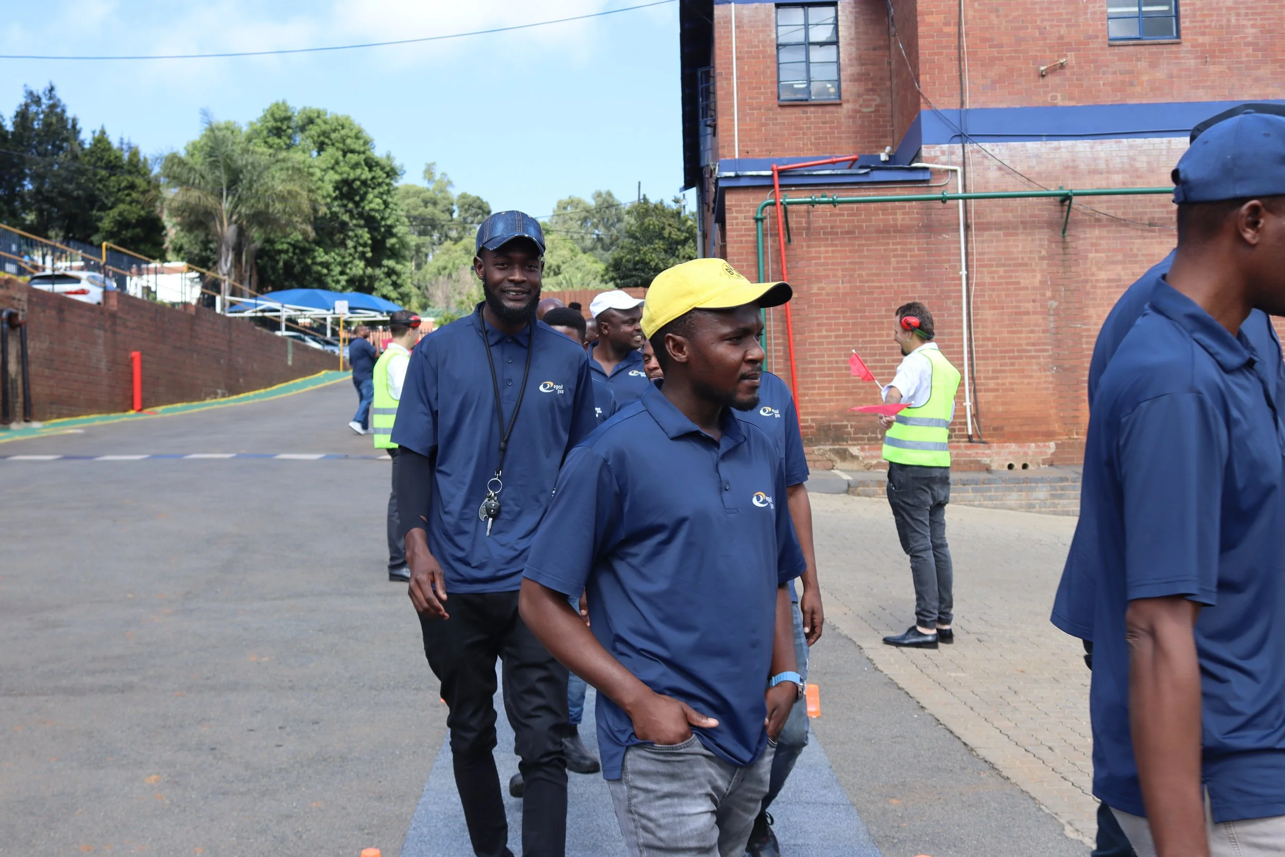 A group of men wearing navy blue shirts and hats walking outdoors near a brick building with trees in the background, some men are smiling and chatting.