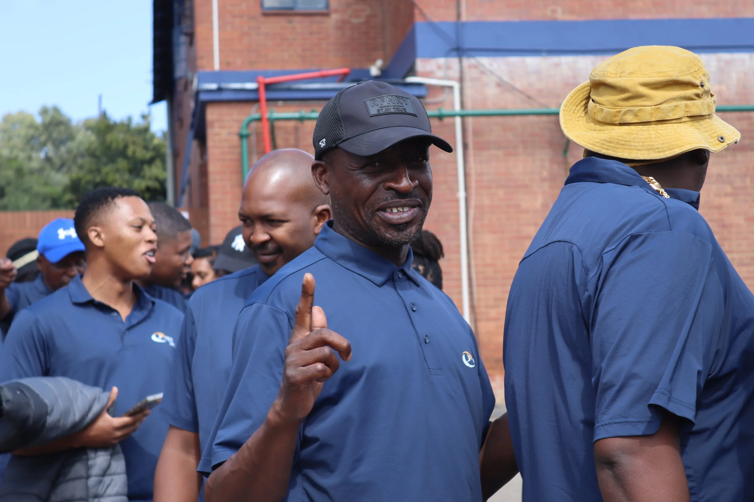 A group of people wearing matching blue shirts outdoors, with some wearing hats. One man in the foreground is giving a thumbs-up. The background includes a brick building and some greenery.