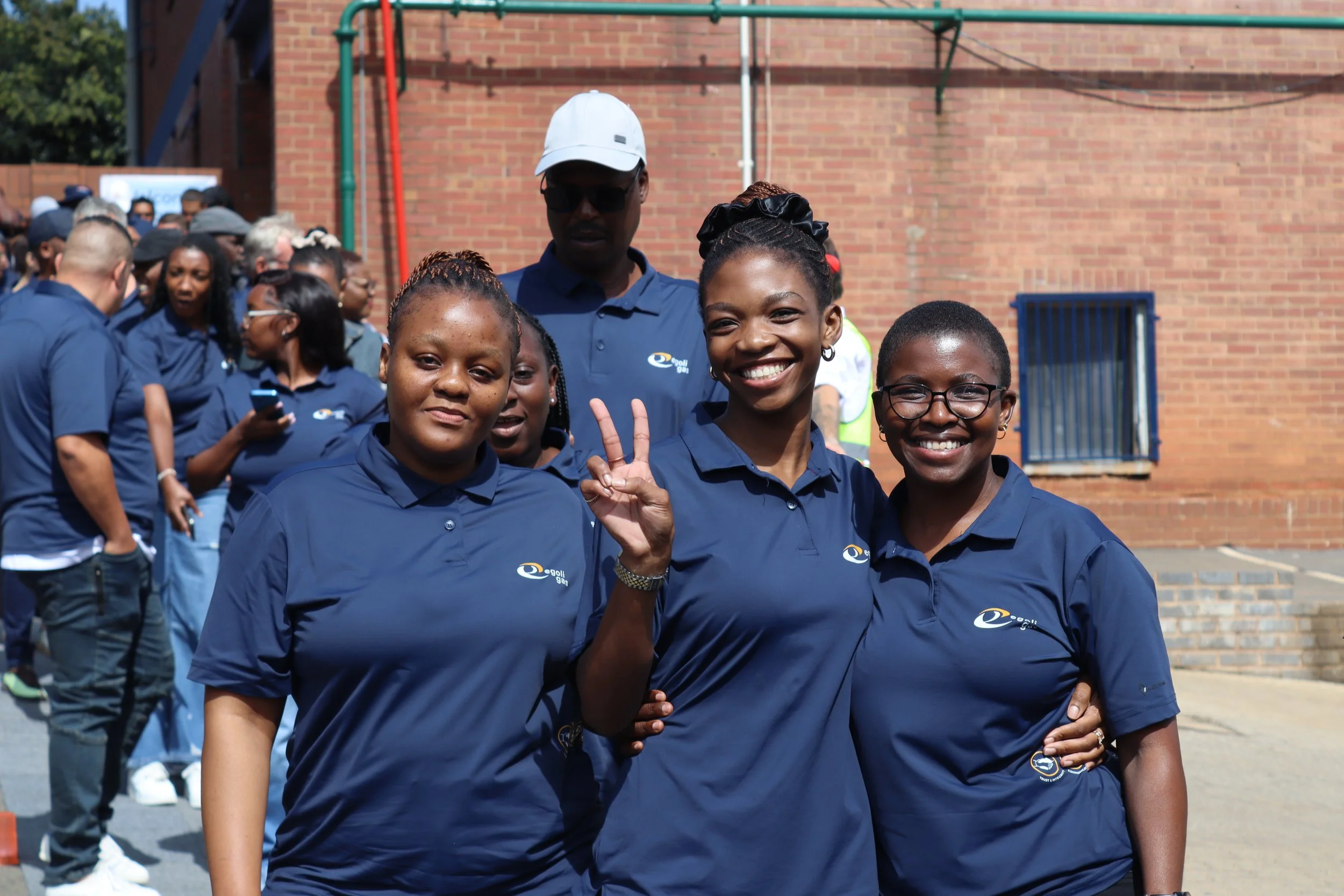 A group of smiling young women and a man in navy blue shirts, with one woman flashing a peace sign, standing outside in front of a brick wall.