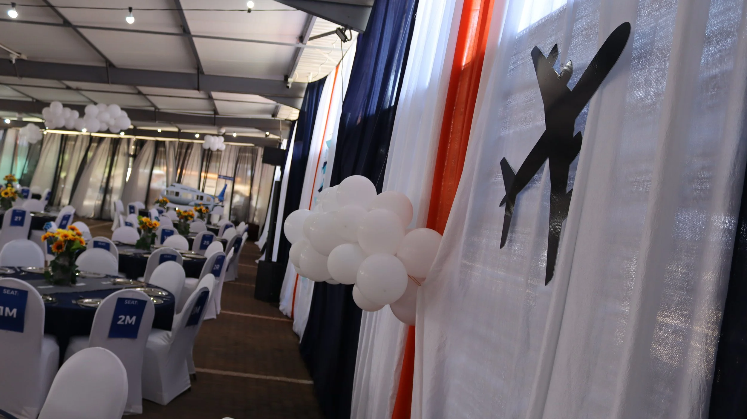 Party or event setup featuring decorated tables with white tablecloths and floral centerpieces, balloons, and a backdrop with black and orange accents, in a spacious venue with draped curtains and ceiling lights.