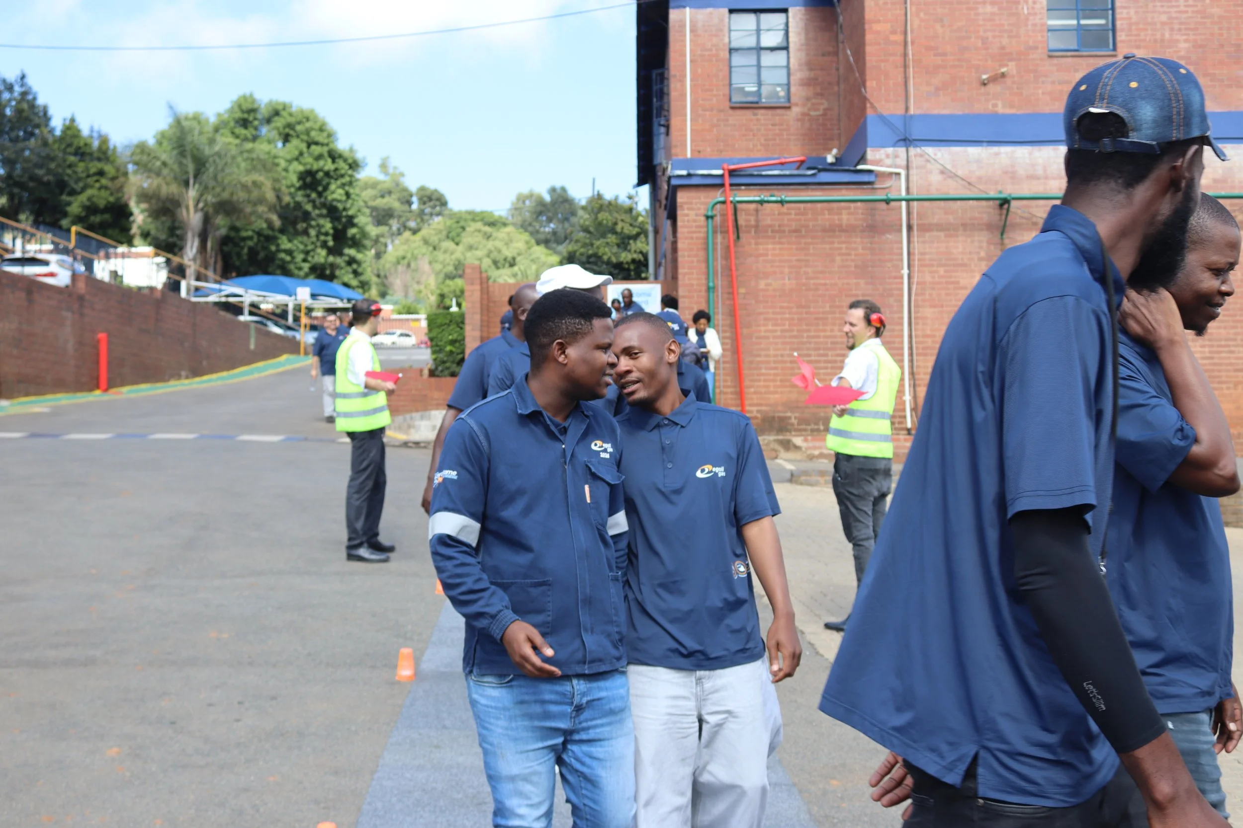 Group of people gathered outside near a brick building, some wearing blue uniforms and yellow safety vests, engaging in conversation and walking.