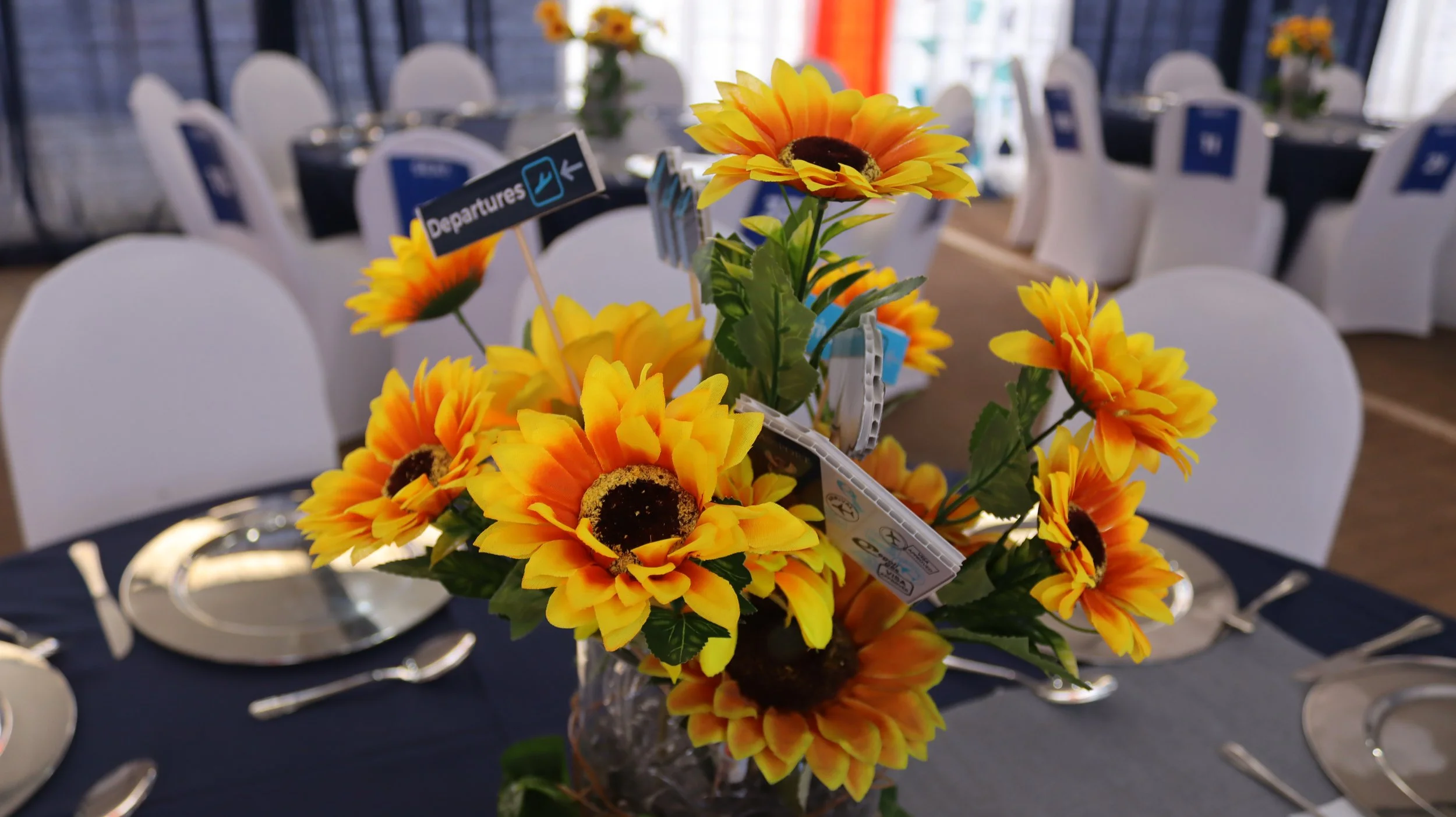 A floral centerpiece with yellow and orange sunflowers on a table set with plates and silverware at an event or banquet.