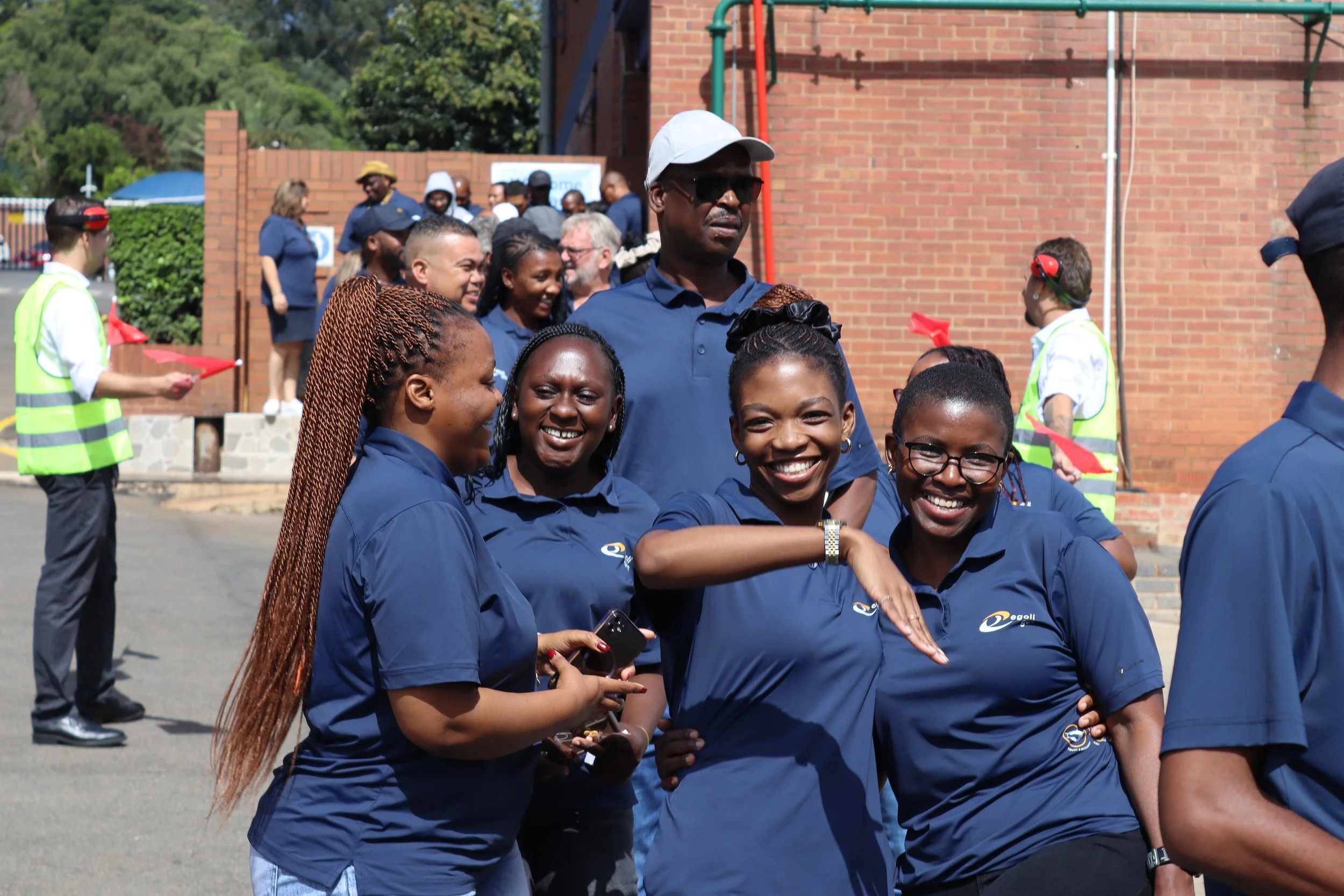 A group of smiling people in blue uniforms gathering outdoors, with some holding phones and engaging in conversations, with a brick wall and trees in the background.