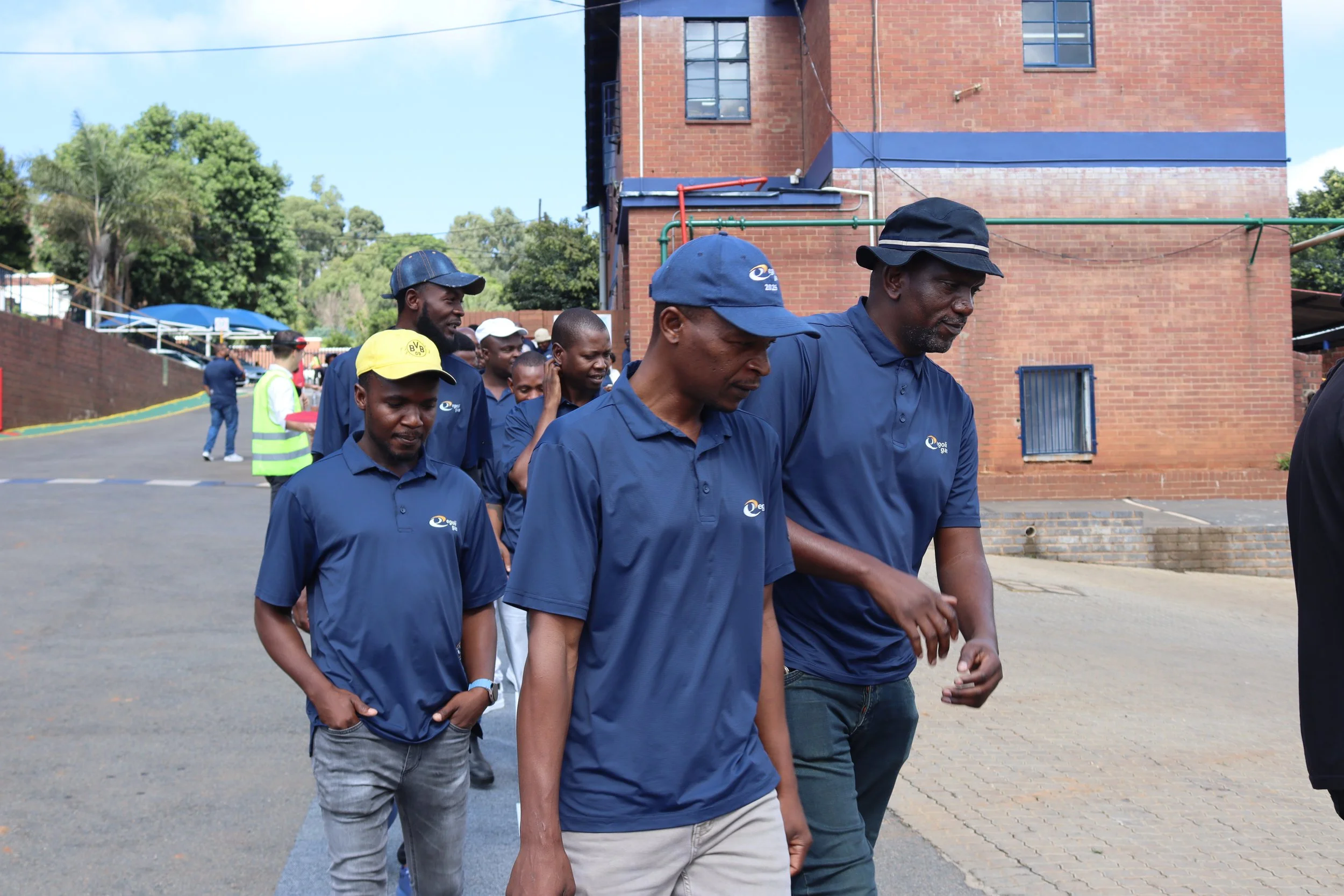 Group of men walking outdoors near a brick building, some wearing matching blue shirts and hats, with greenery and a blue sky in the background.