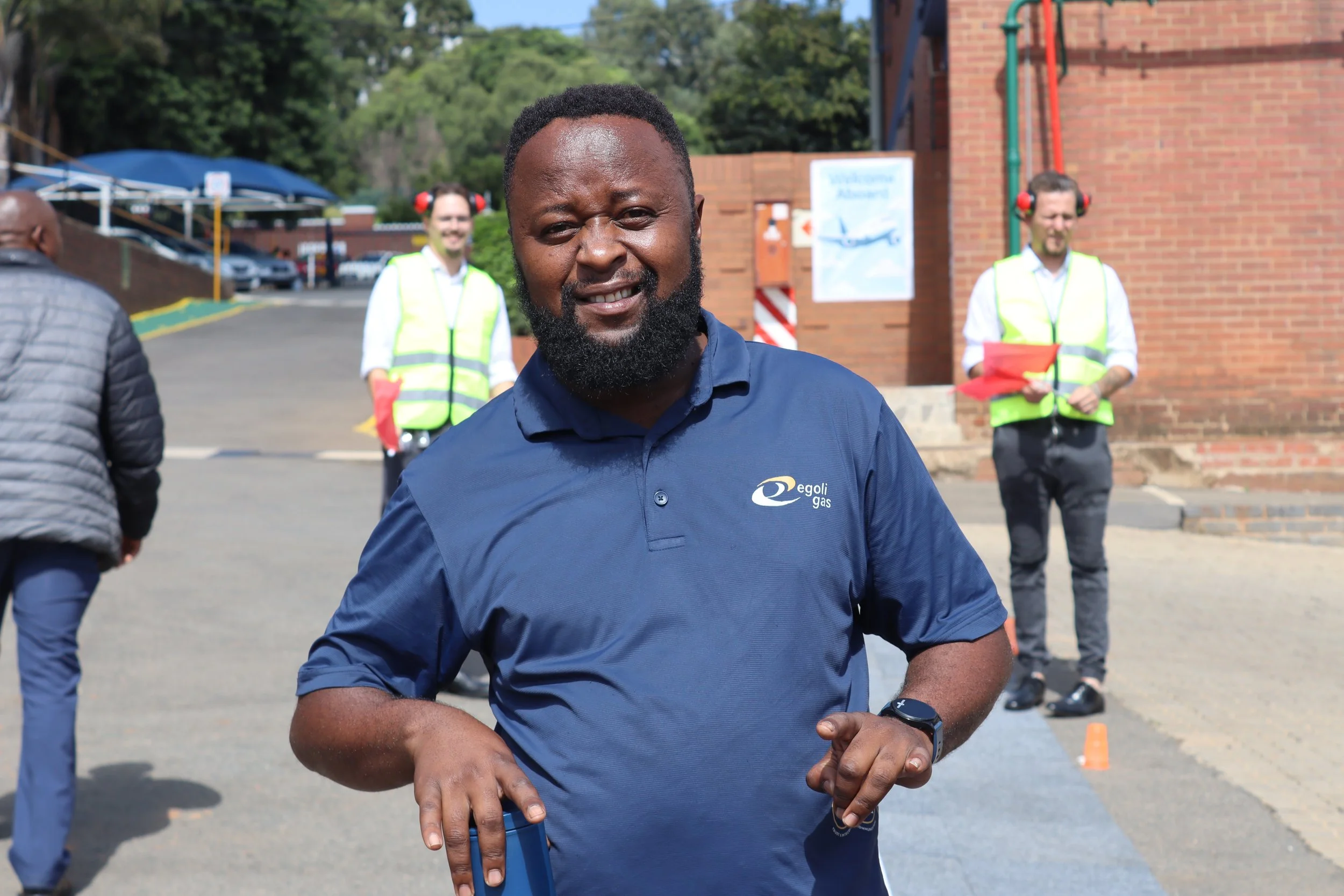 A smiling man with a beard in a navy blue shirt with a company logo on it, standing outdoors during daytime with other people in the background, some wearing yellow safety vests and ear protection, in front of a brick building and trees.