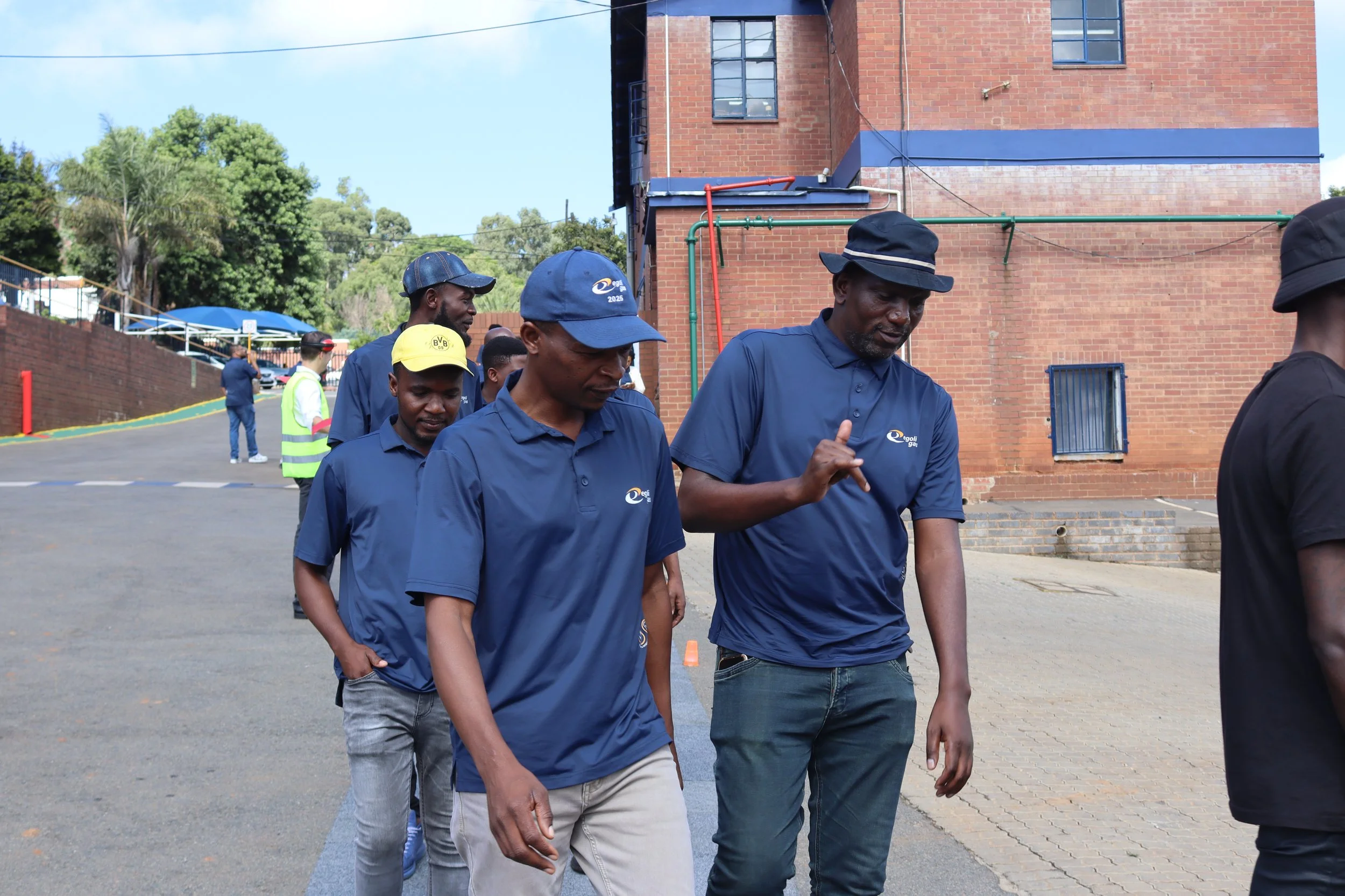 A group of men walking outdoors in front of a brick building, with some greenery and trees in the background. The men are wearing blue and black clothing, with some sporting caps.