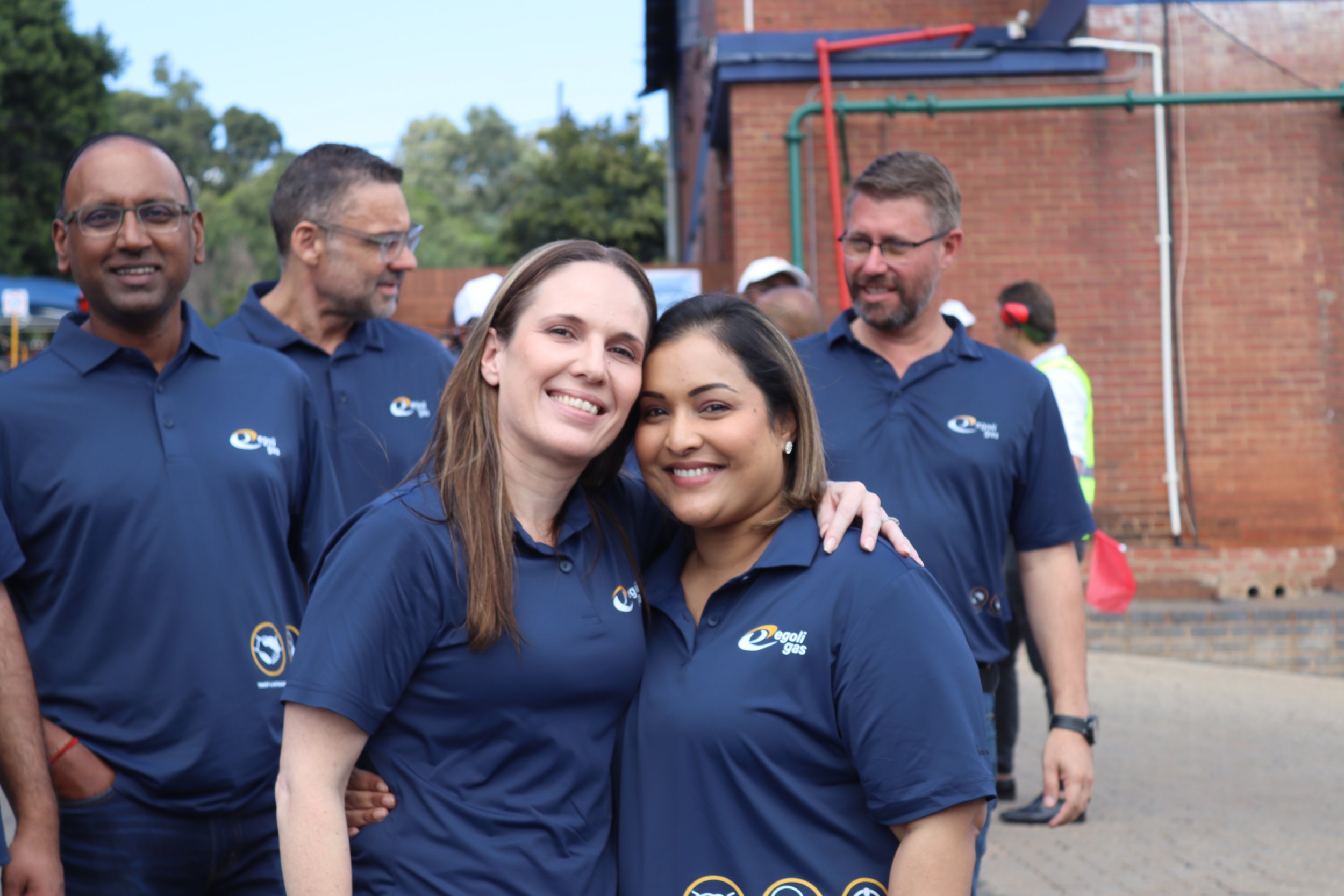 A group of six diverse adults, wearing matching blue shirts with a logo, are gathered outdoors at a community event, smiling and standing close together.