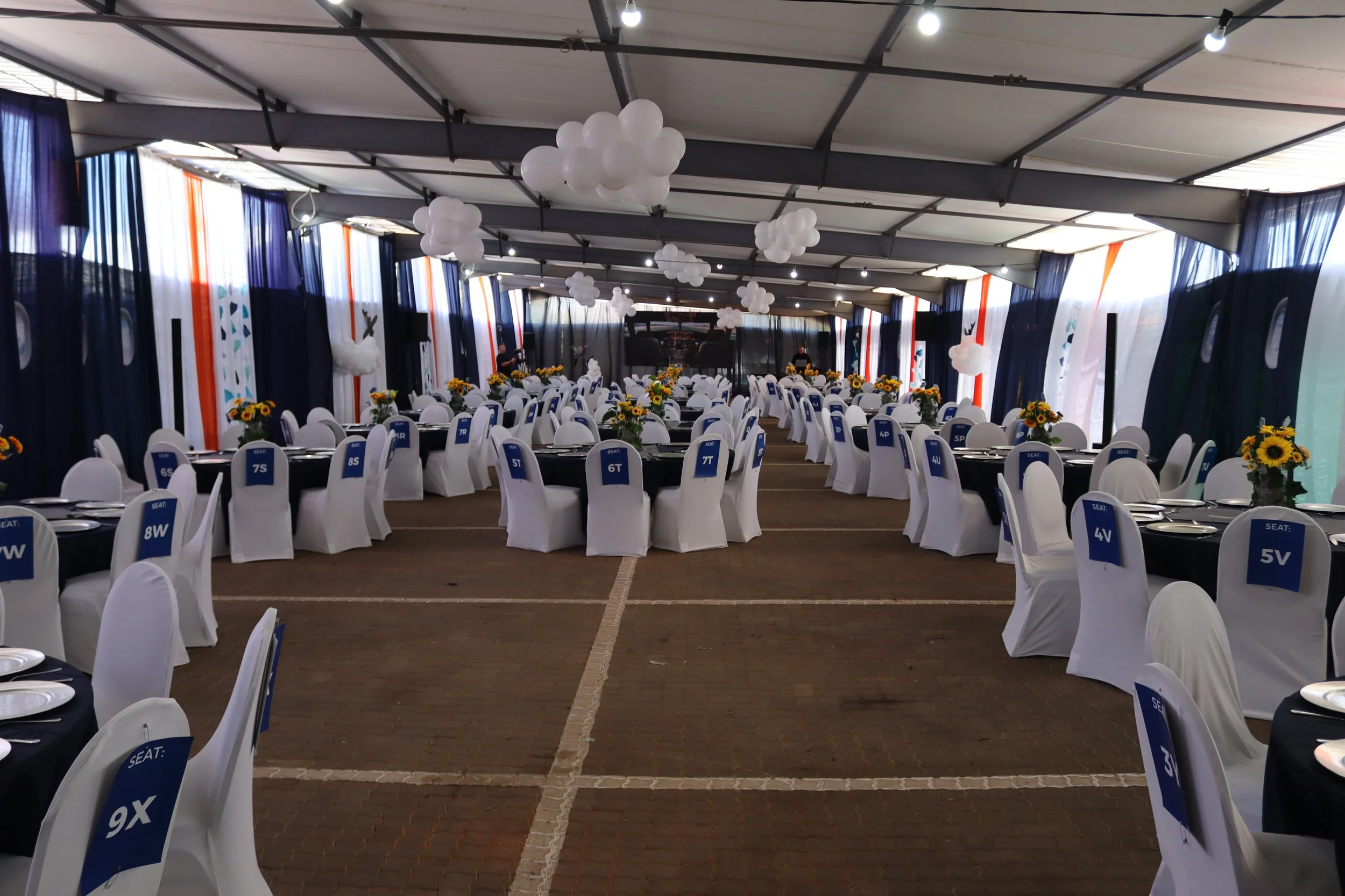 A large event tent decorated with white balloons hanging from the ceiling, dark blue and white curtains along the sides, and round tables with white chairs labeled with seat numbers, set for a formal event.