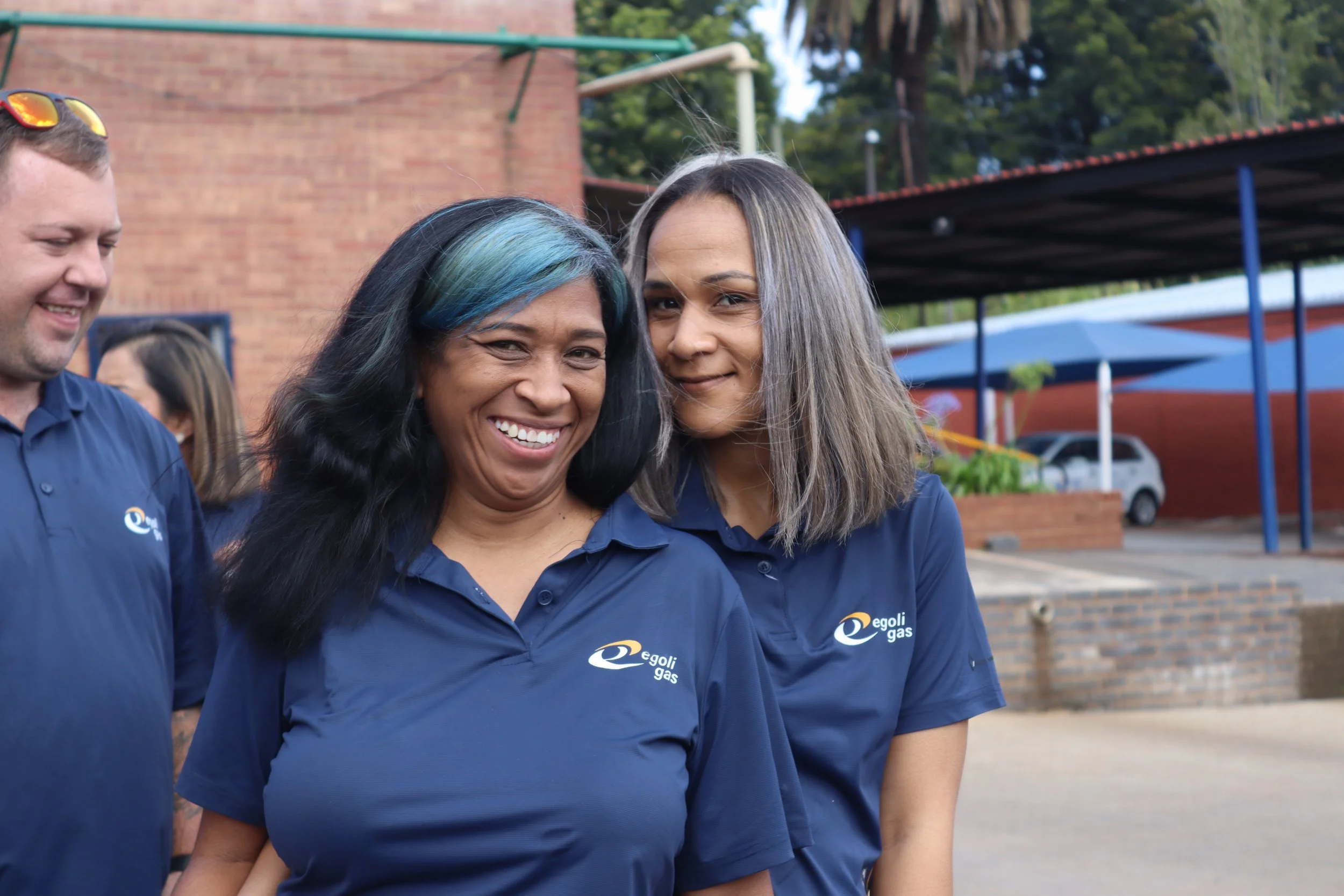 Three women and one man standing outdoors, wearing matching blue polo shirts with 'Egoli Gas' logos, smiling, with a brick wall, trees, and a parking lot in the background.