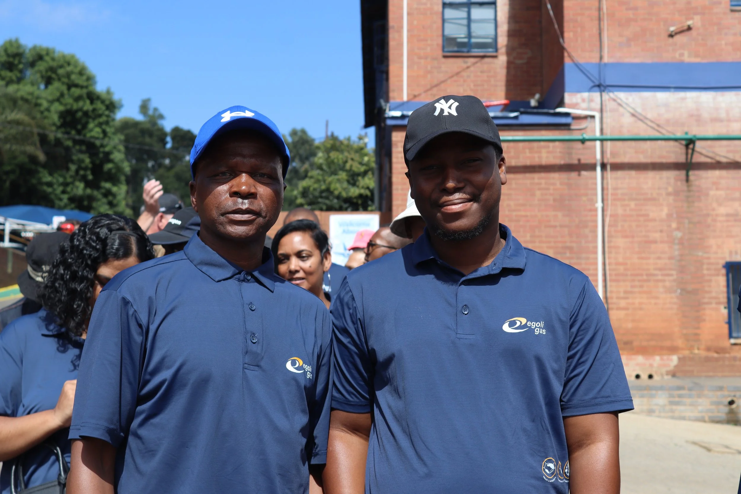 Two men wearing blue shirts and caps standing outdoors in front of a brick building and trees, with a crowd of people in the background.