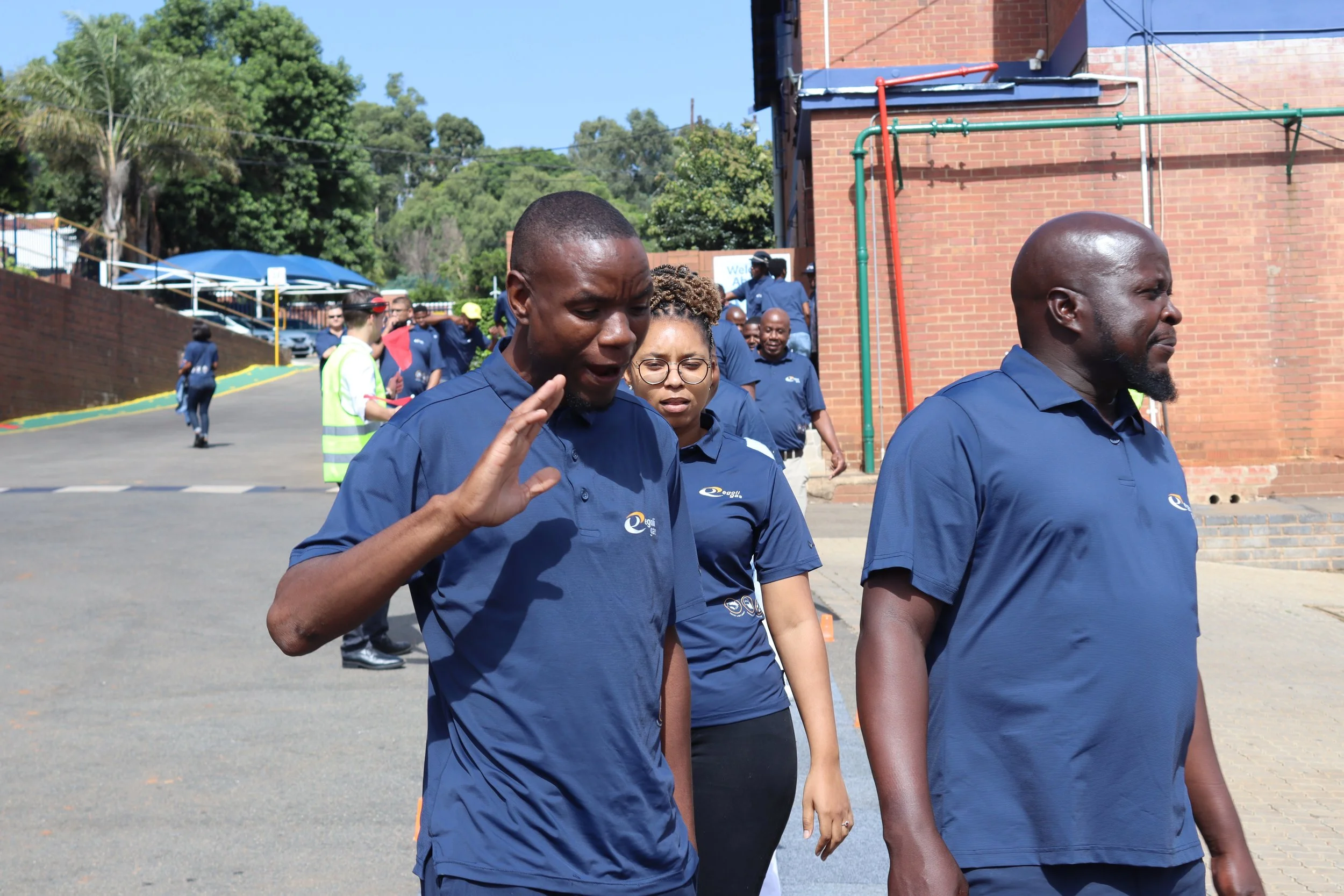A group of people walking outdoors, including a man with a shaved head and beard, a woman with glasses and curly hair, and others in matching blue shirts.