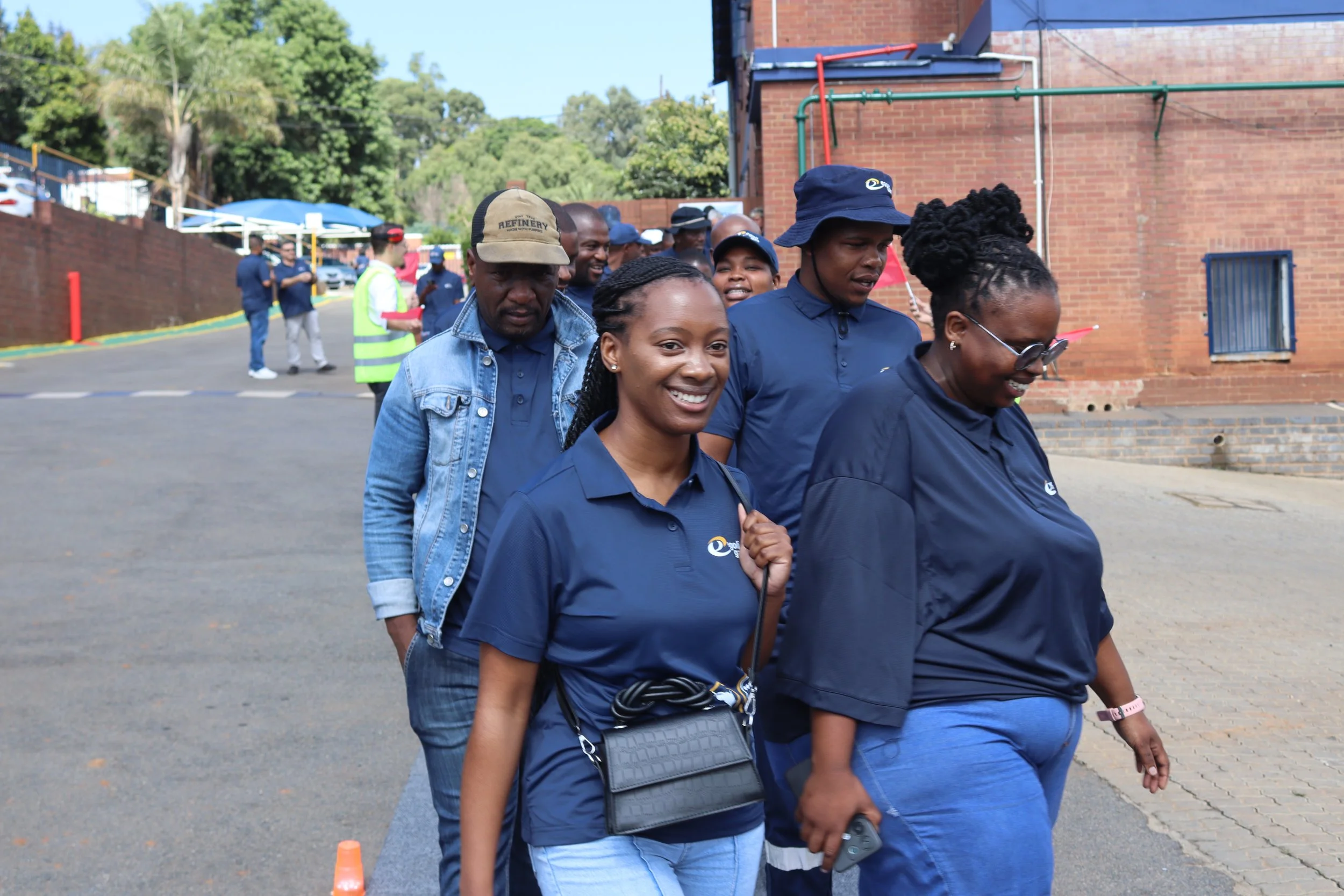 A group of diverse people walking outdoors on a sunny day, near a brick building with trees in the background, some smiling and engaging with each other.