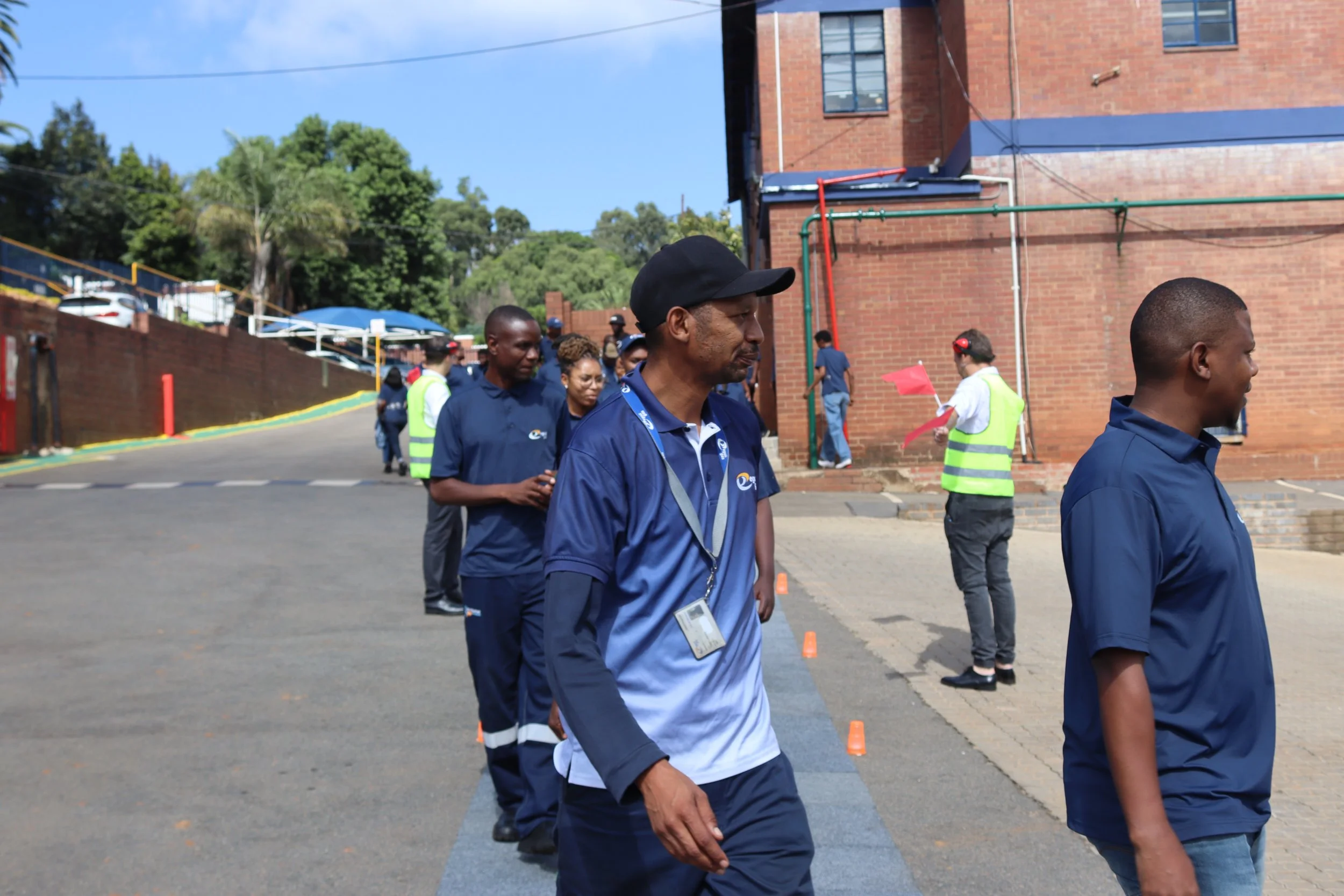 A group of people, including men and women, walking outdoors near a brick building, with some wearing lanyards and safety vests.