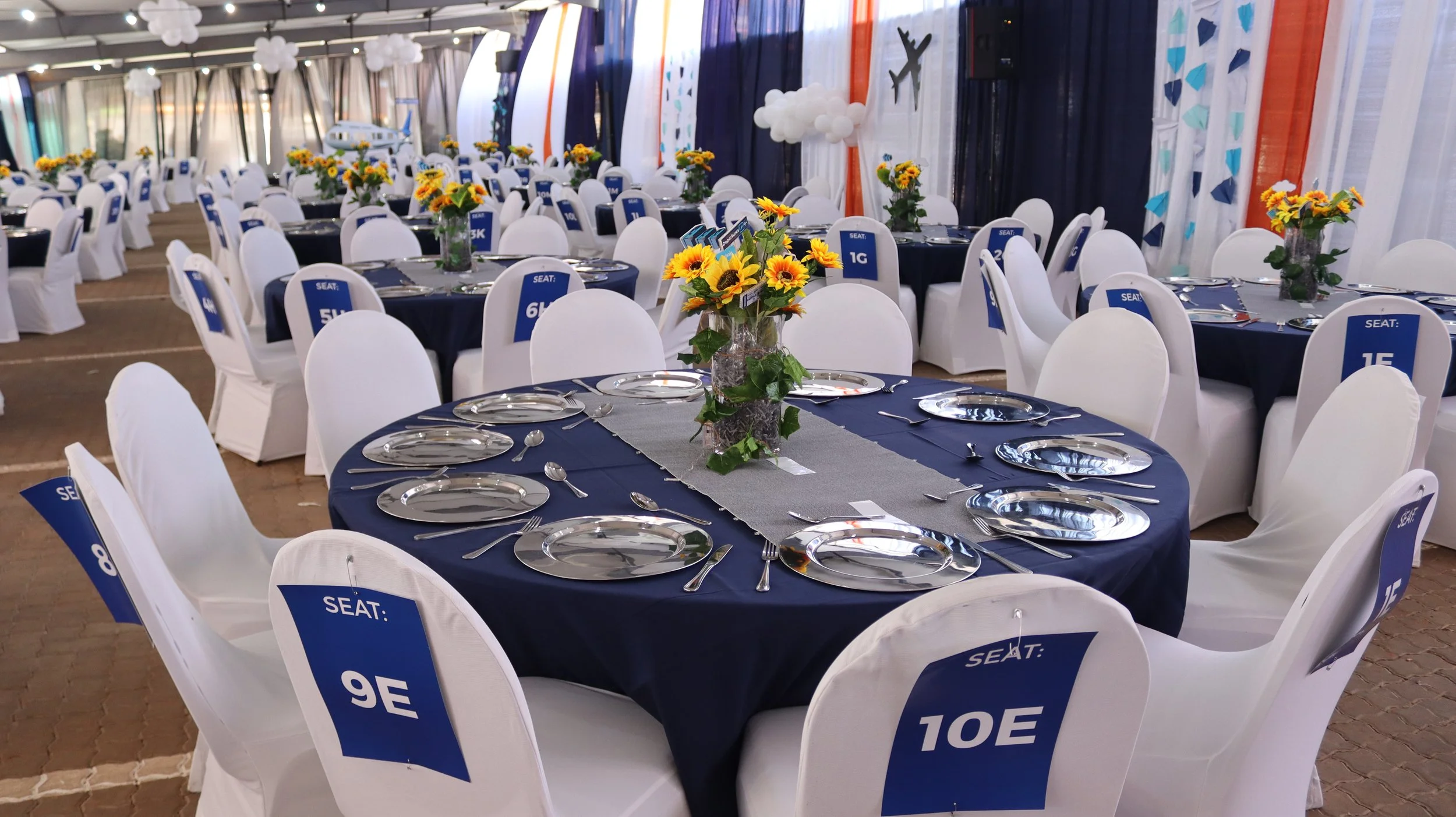 Event space decorated with round tables covered with navy blue tablecloths and white chairs with seat number signs, floral centerpieces with sunflowers, and airplane-themed wall decorations.