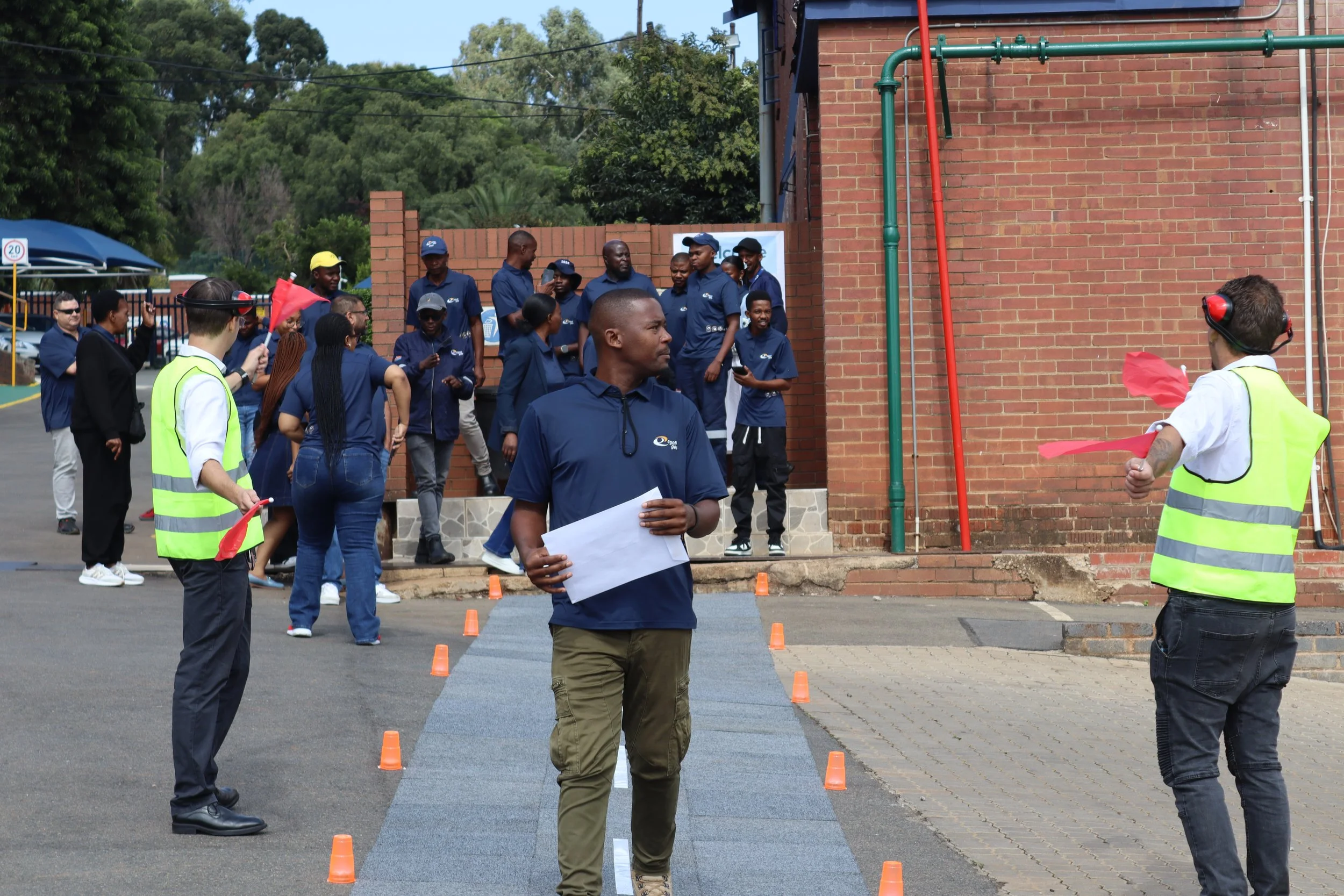 A group of people gathered outside near a brick building, some holding red flags and wearing yellow safety vests, appearing to participate in a demonstration or event involving traffic cones and a blue walkway.