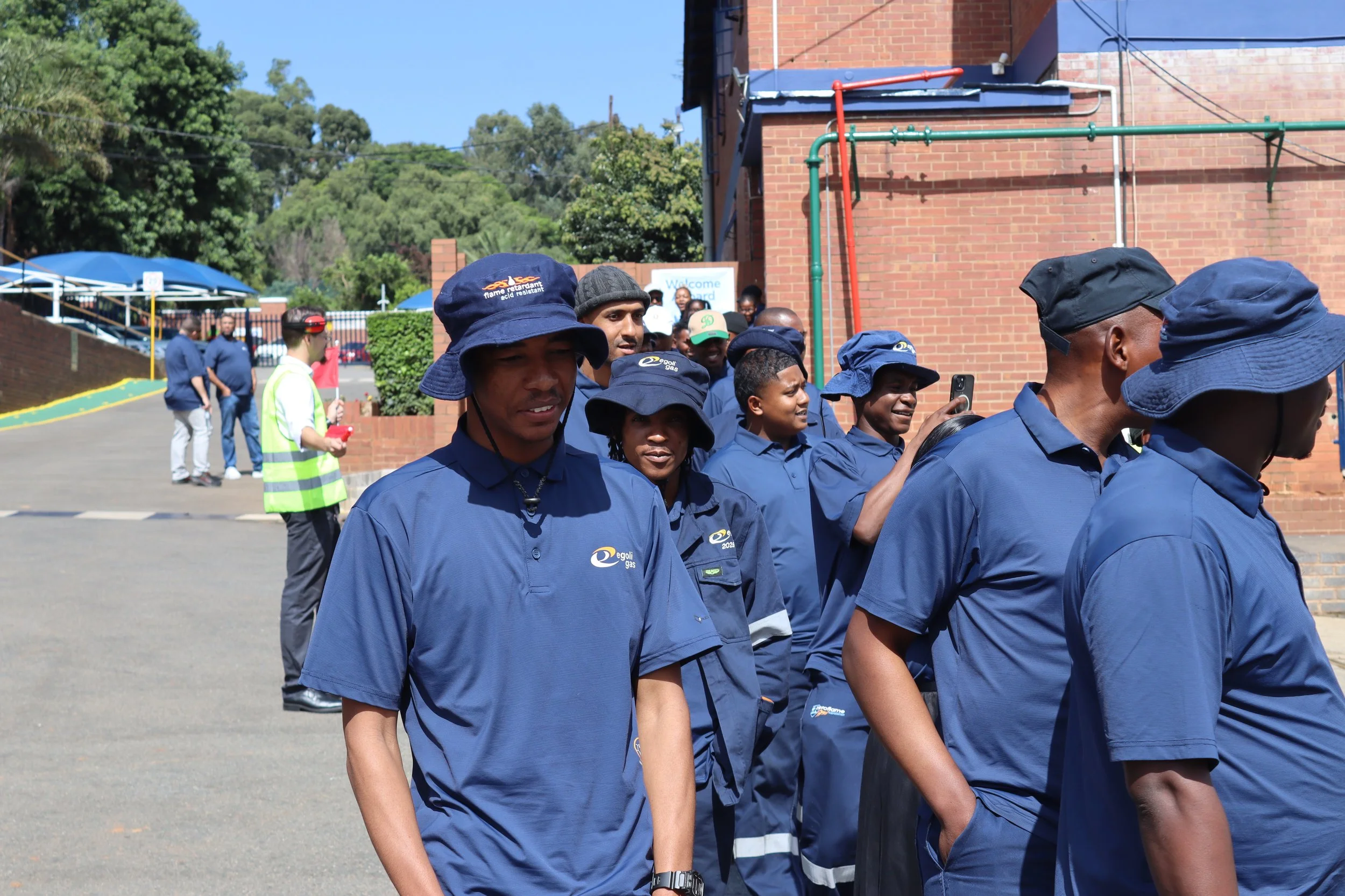 A group of people in blue uniforms and hats standing outdoors near a brick building, with trees and a parking lot in the background.