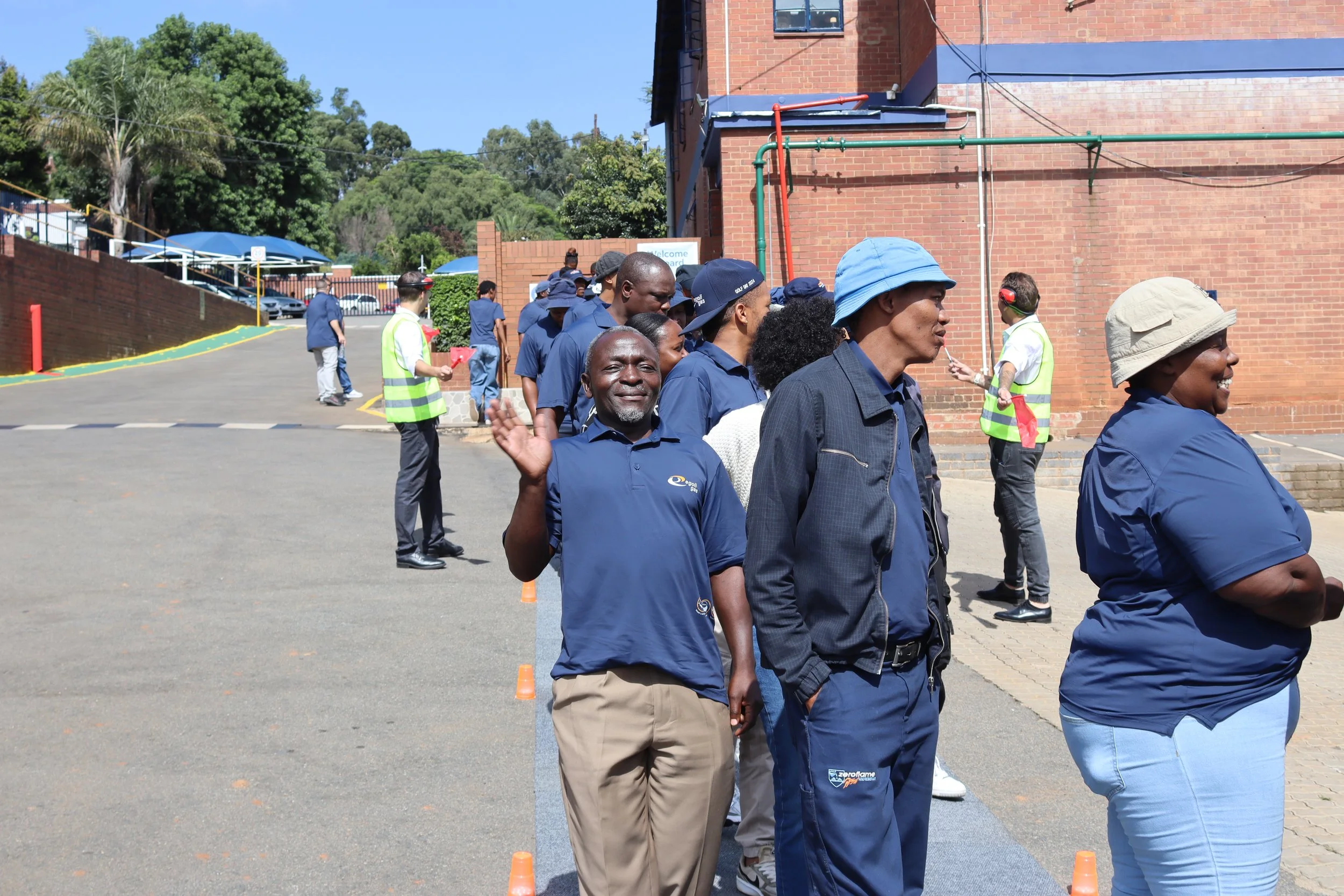 A group of people standing in line outdoors, some wearing blue uniforms and hats, with orange cones on the ground marking the line, in front of a brick building and surrounded by greenery.