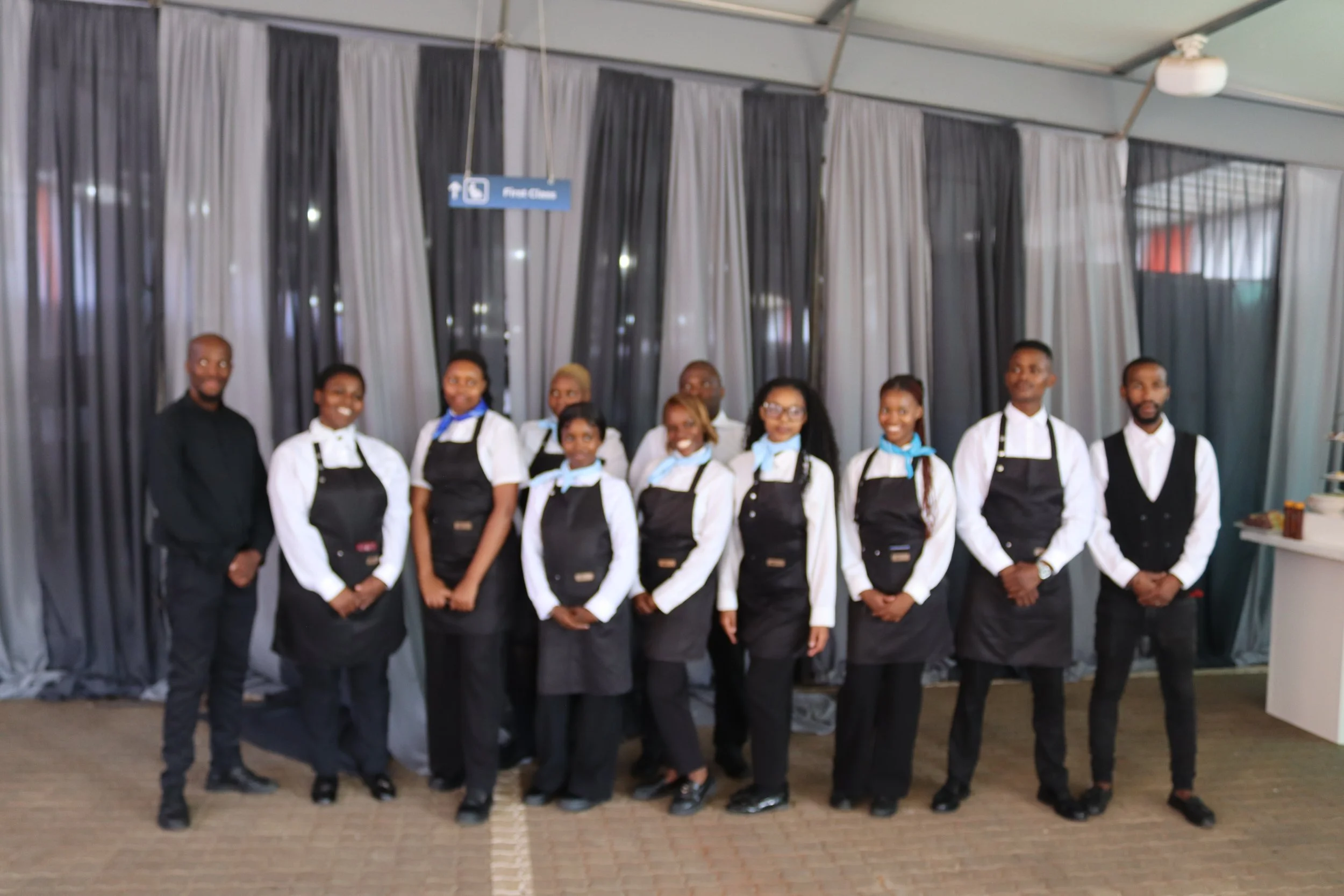 Group of nine people standing in a row indoors, dressed in formal white shirts and black aprons, with some wearing light blue scarves or bow ties, in front of black and white curtains. They are smiling and posing for the photo.