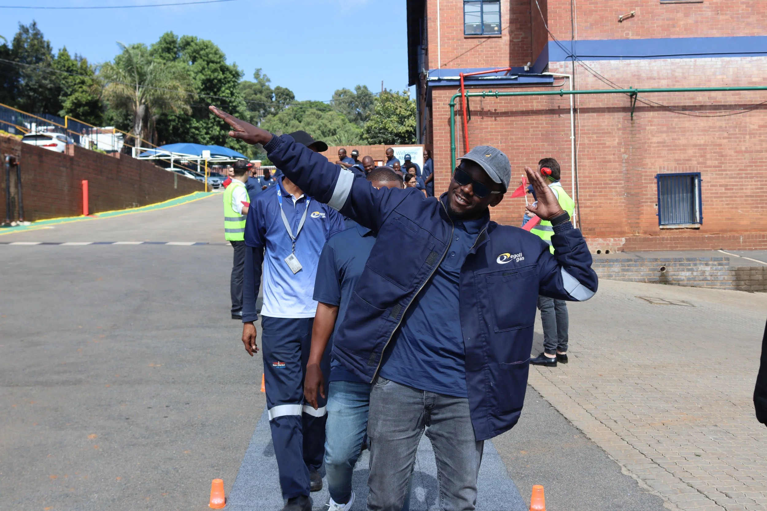 A man wearing dark sunglasses, a gray bucket hat, and a navy jacket smiling and giving a playful salute with his hand. He is walking outdoors on a paved area with small orange cones placed on the ground and a group of people in the background near a 