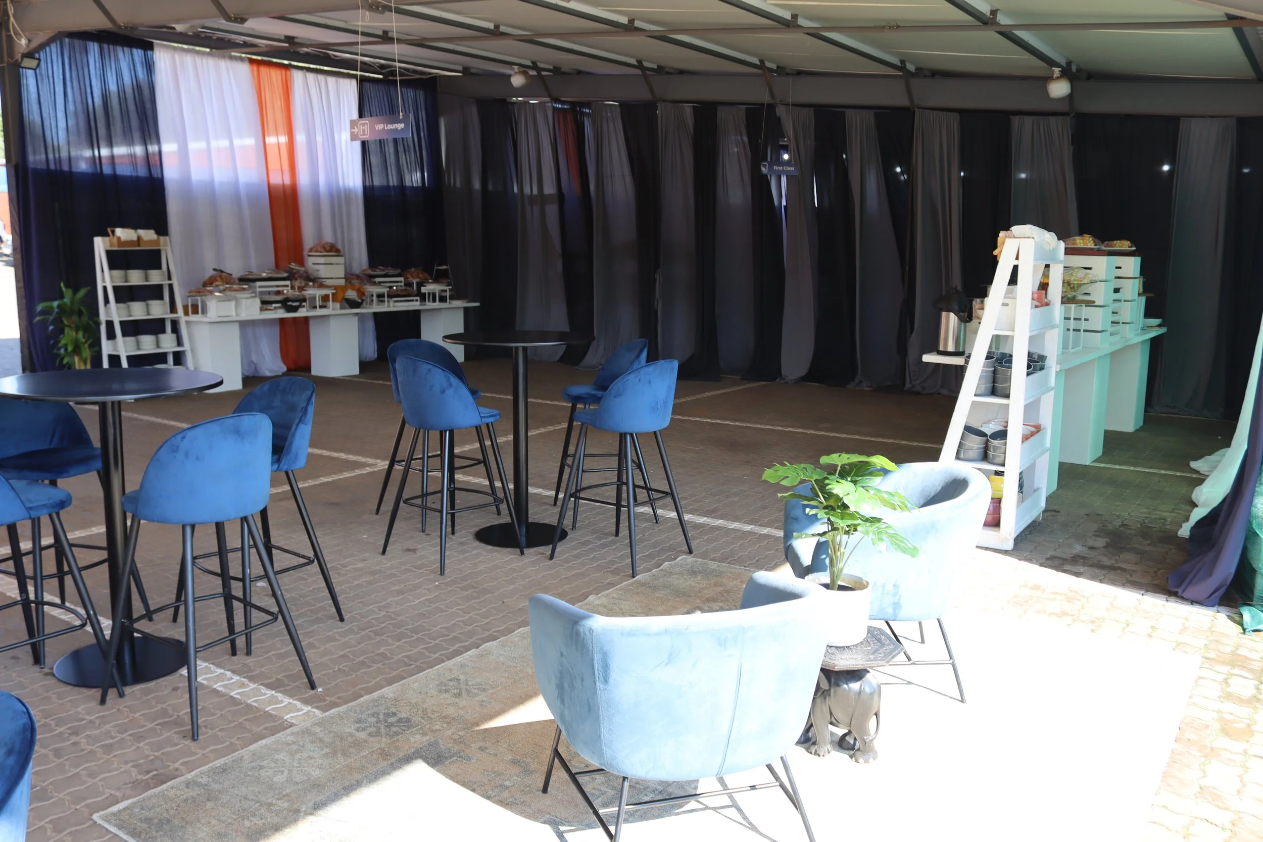 A buffet setup inside a tent with tables of food and seating, including blue chairs, black tables, and white shelves with dishes and food containers.